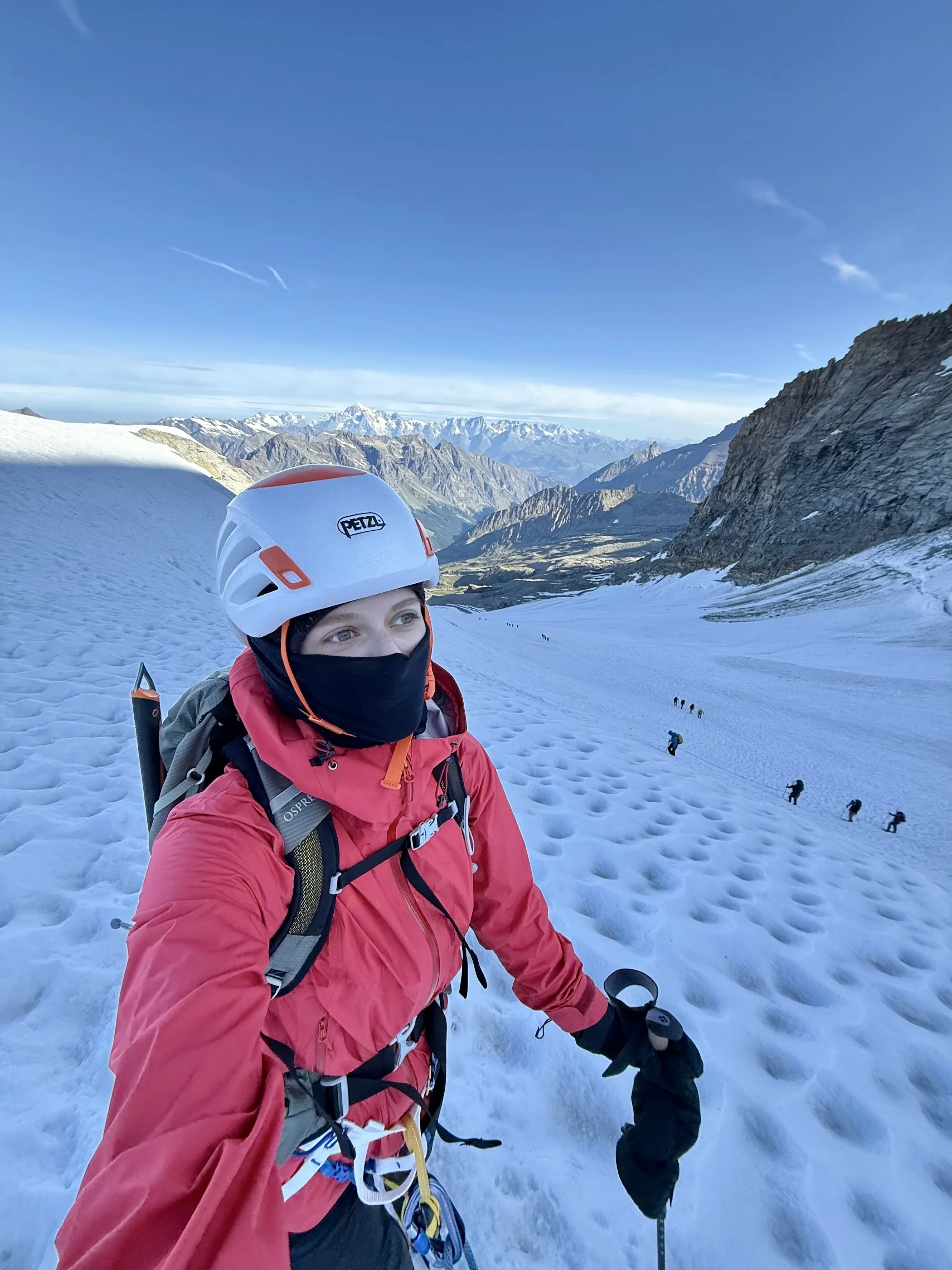 A mountain climber dressed in red gear and a white helmet stands on snowy terrain with a mountain range and blue sky in the background.