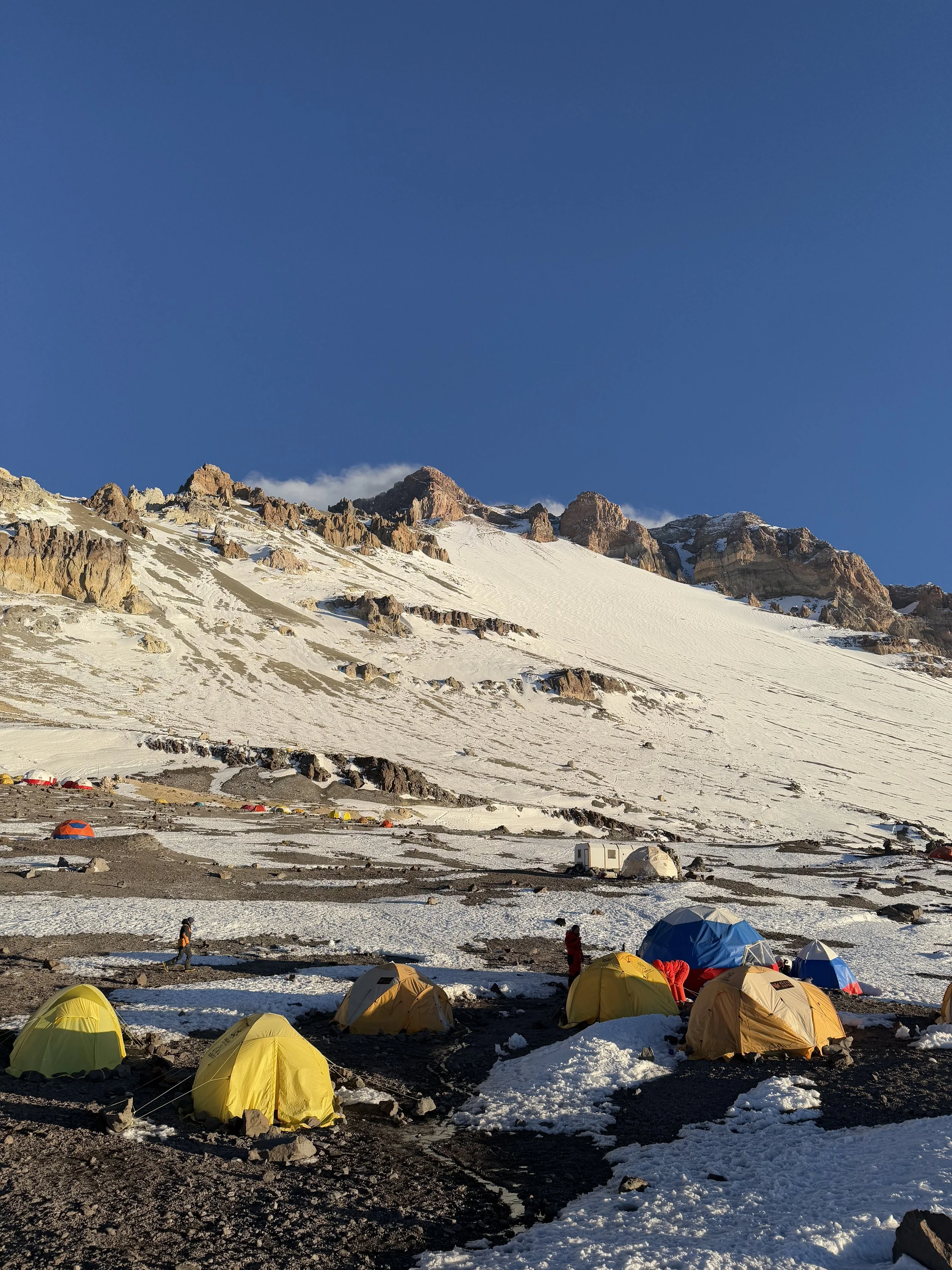 Camping tents on snow at the base of a mountain with rocky peaks and a clear blue sky.