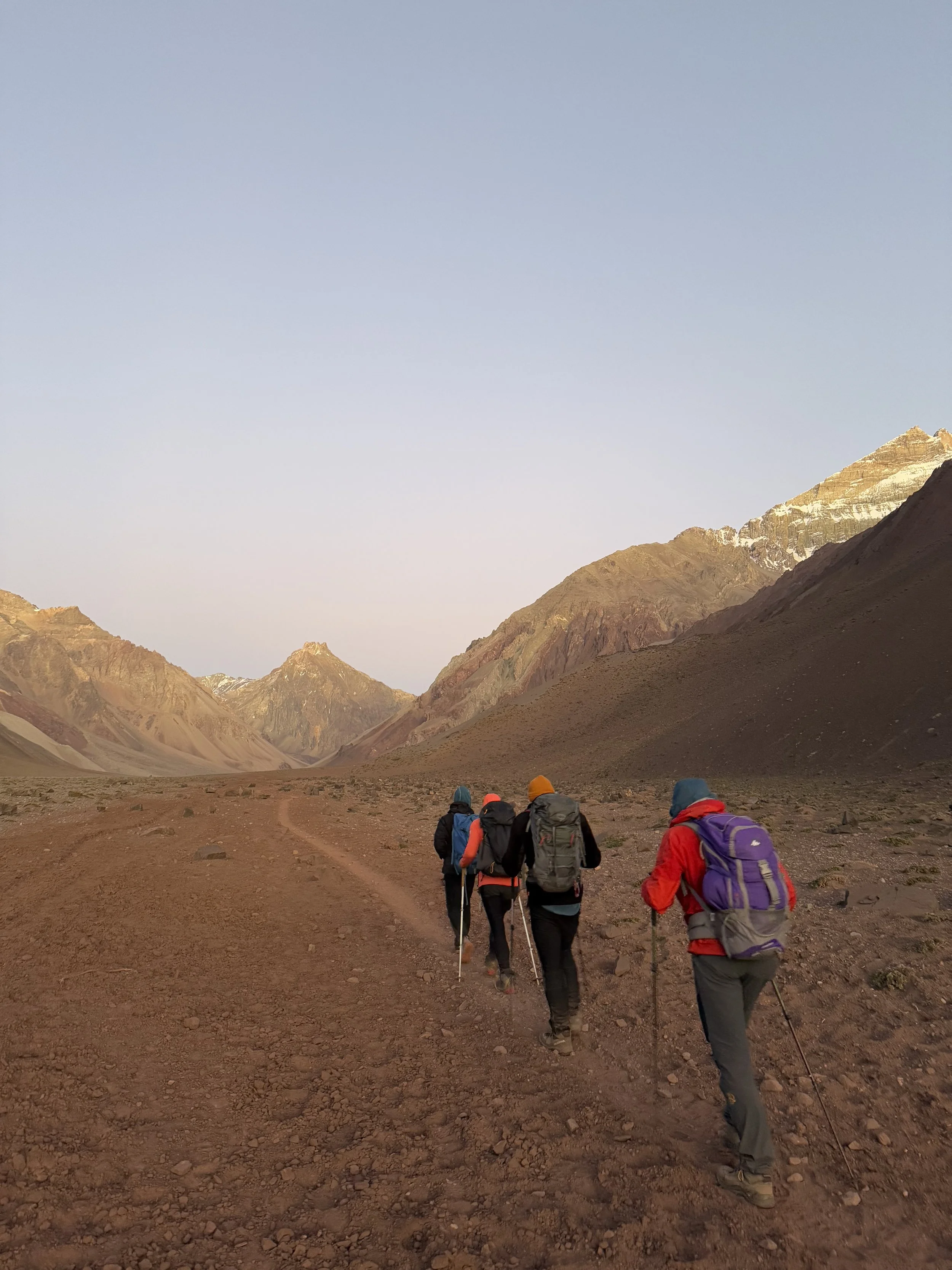 Five hikers with backpacks walking on a dirt trail through a mountainous desert landscape at dawn or dusk.