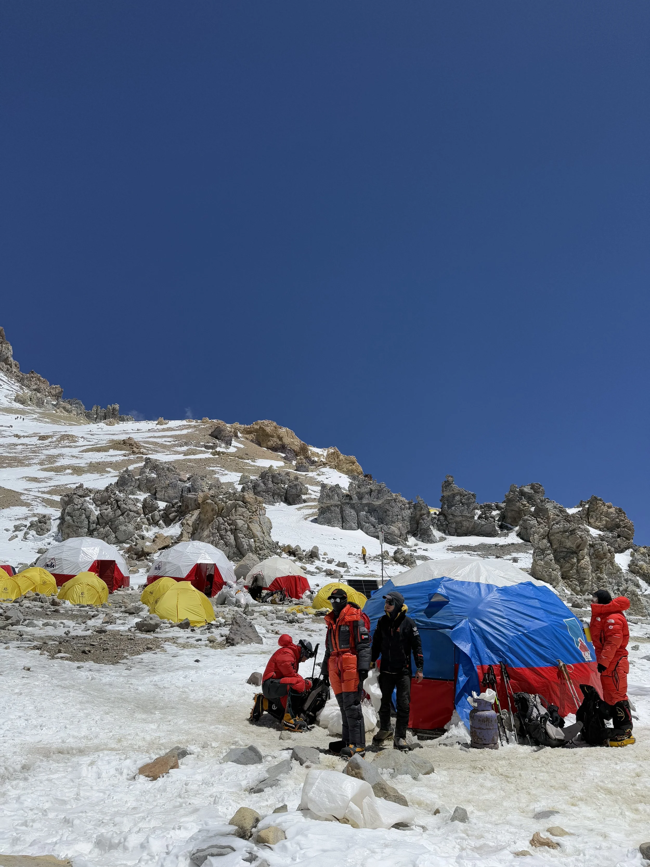 Mountaineers in warm gear setting up a campsite with tents on snow-covered mountain, rocks, and a clear blue sky.