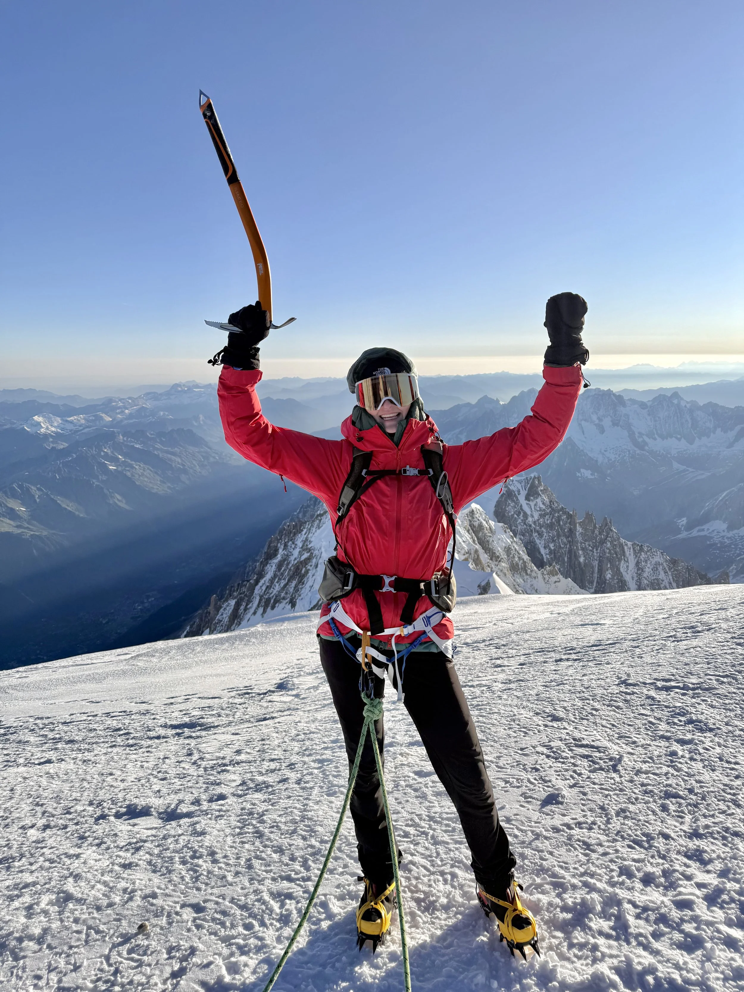 Person in red mountaineering gear celebrating on snow-covered mountain peak with mountain range in background.