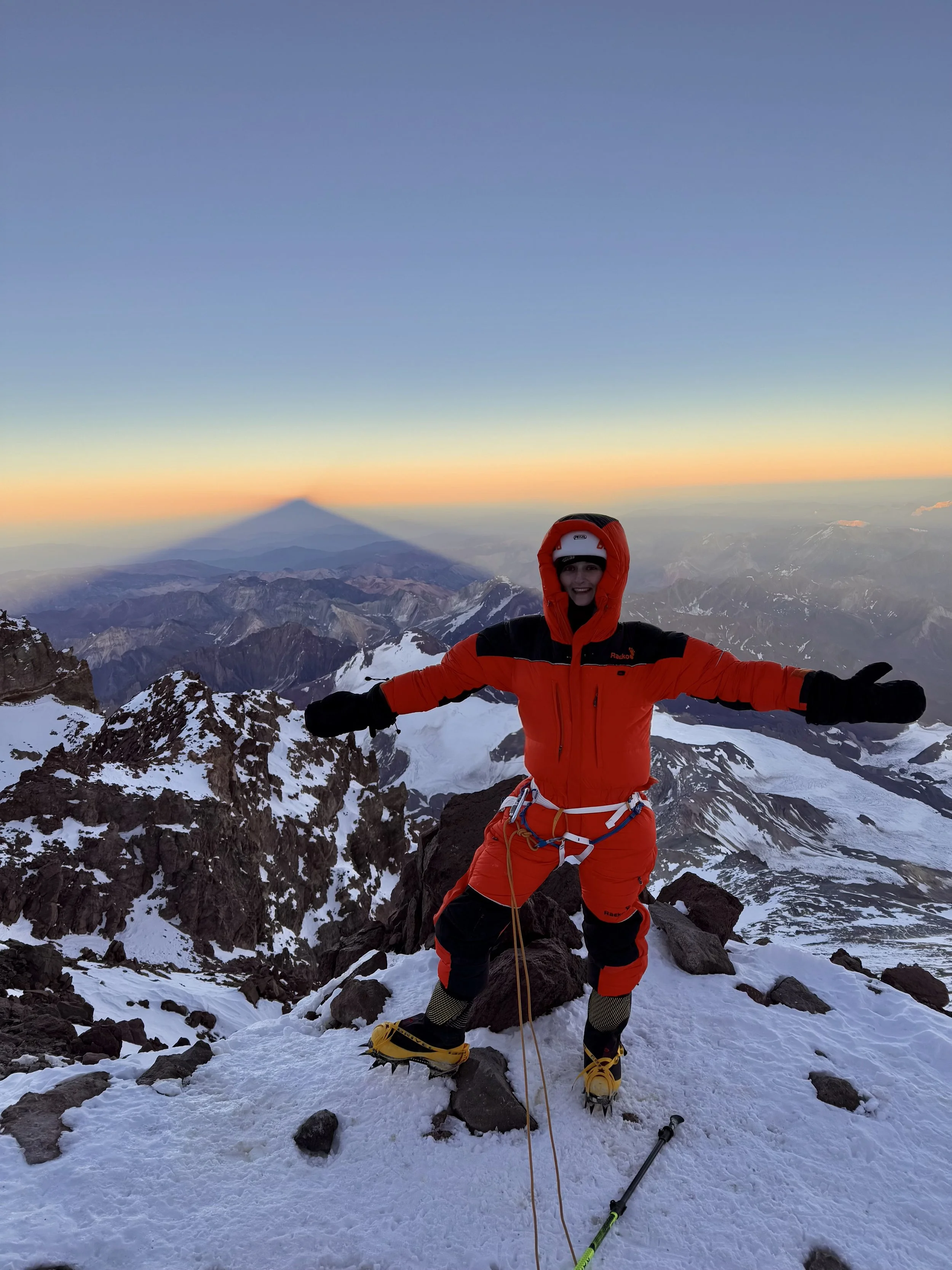A person dressed in red mountaineering gear standing triumphantly on snowy mountain peak during sunset, with snow-capped mountains in the background and the shadow of a volcano on the horizon.