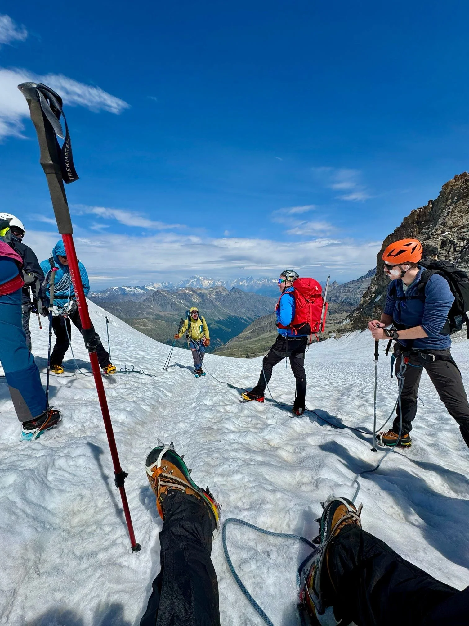 Group of hikers standing on snow-covered mountain slope with trekking poles, wearing helmets, backpacks, and winter gear, with a scenic mountainous landscape in the background.