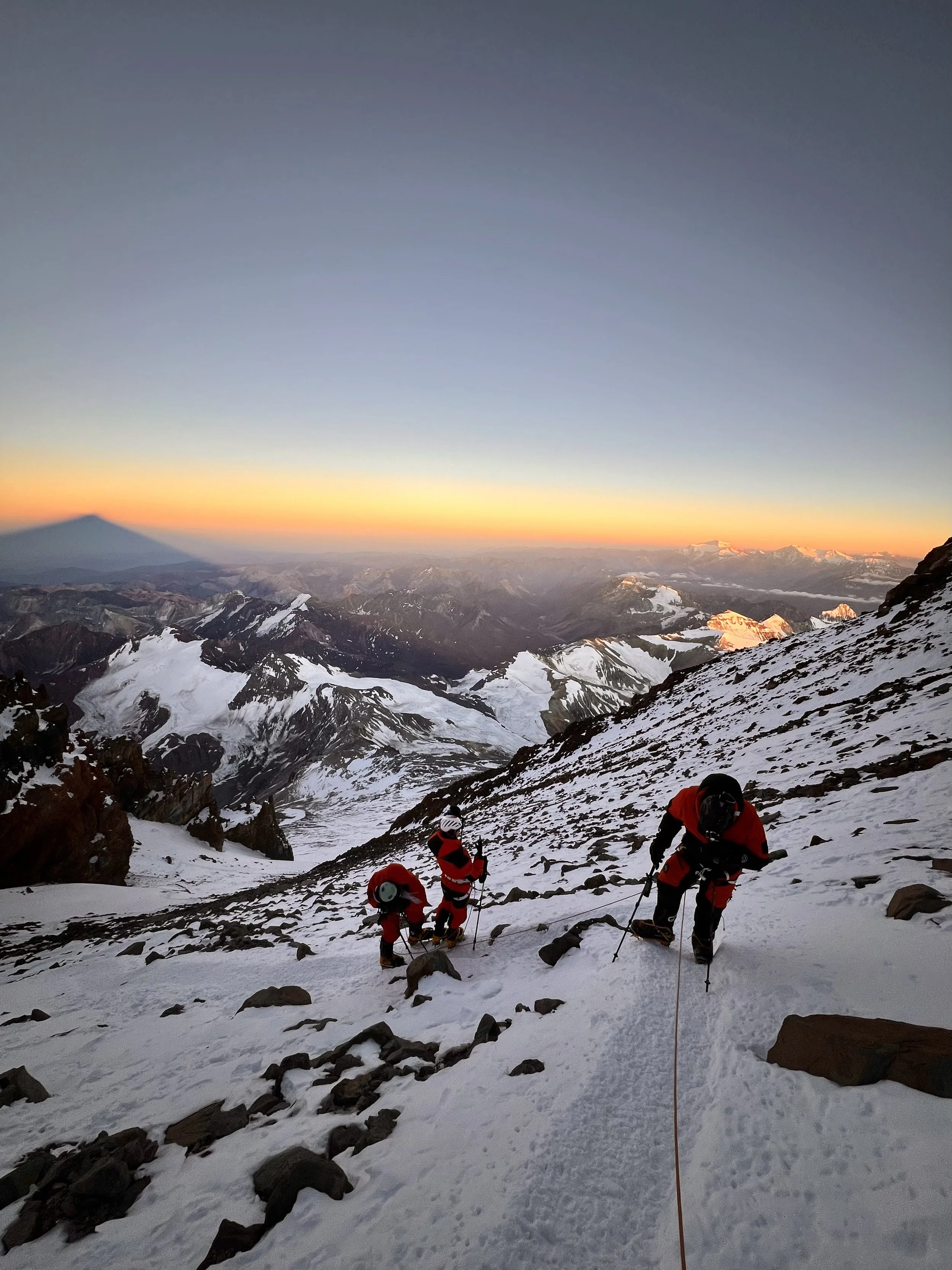 Three mountaineers climbing a snowy and rocky mountain slope at sunrise, with a panoramic view of snow-capped peaks and a colorful sky in the background.