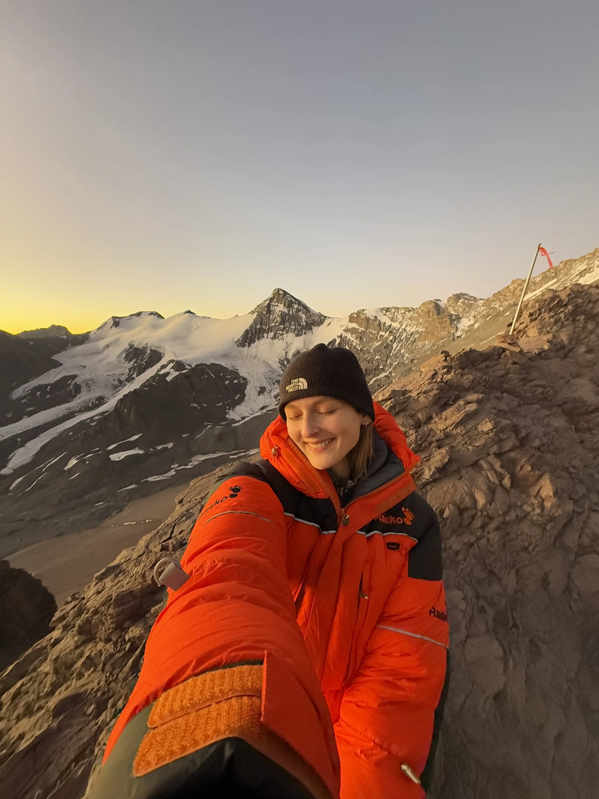 A smiling woman dressed in an orange and black outdoor jacket and a black beanie taking a selfie on a rocky mountain slope during sunset, with snow-covered peaks in the background.