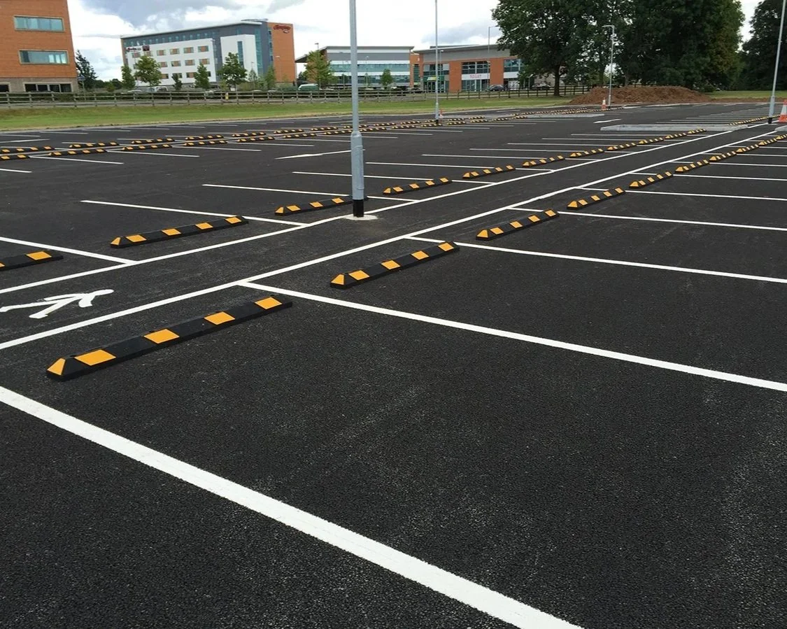 Empty parking lot with newly painted white lines, parking blocks, a designated handicapped space, and some lamp posts. Buildings and trees are visible in the background.