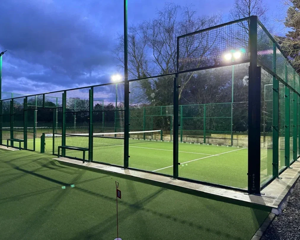 An illuminated padel tennis court surrounded by high green fences, with a net in the center and tall floodlights overhead, set during dusk with cloudy sky and leafless trees in the background.