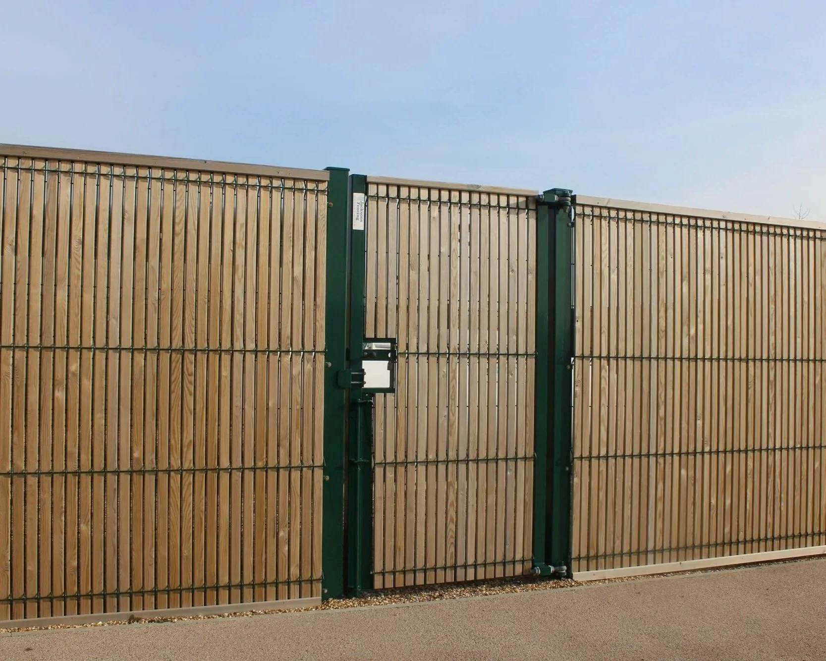 A tall wooden fence with a green metal gate and gate opening mechanism, under a clear blue sky.