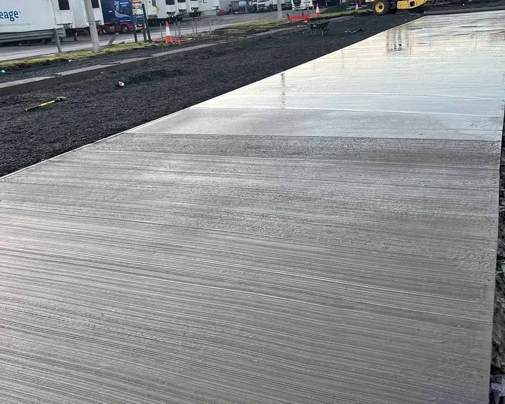 Freshly poured concrete sidewalk with visible trowel marks, adjacent to an area of dark soil and construction tools, with parked trucks in the background.