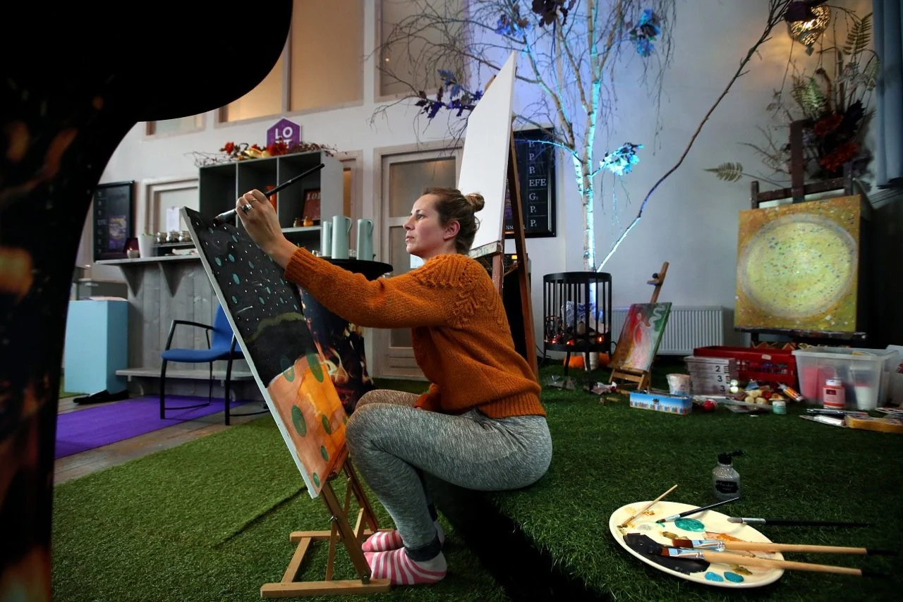 A woman sitting cross-legged on artificial grass, painting a colorful landscape on a canvas in an indoor art studio decorated with trees, paintings, and art supplies.