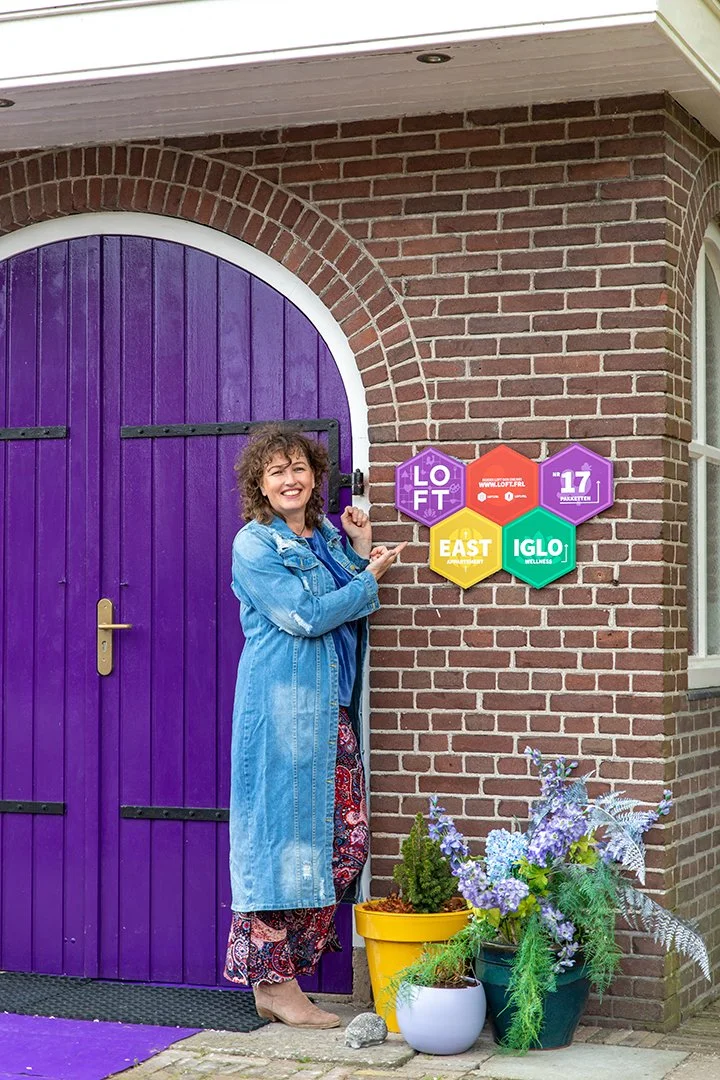 Woman standing outside near a purple door with colorful hexagon signs on brick wall, plants in pots on ground.