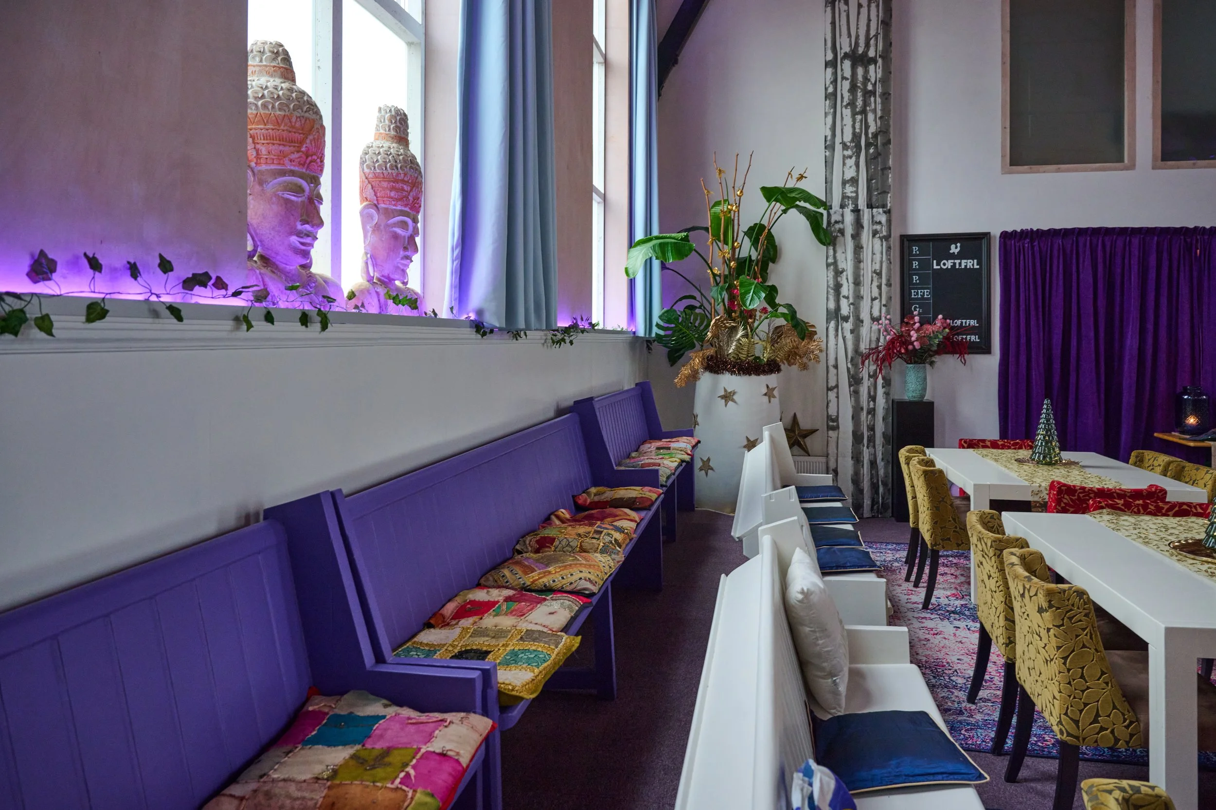 Interior of a colorful dining area with purple benches, white chairs, and patterned cushions, decorated with plants, fairy lights, and themed decorations like stars and a menu board in the background.