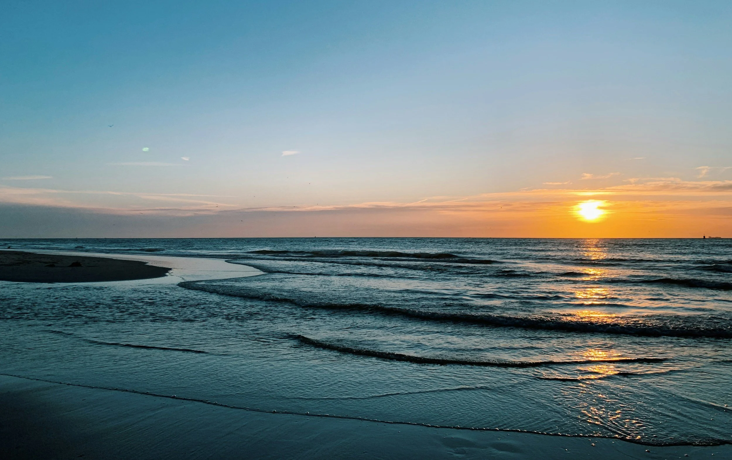 Sunset over the ocean with gentle waves and a sandy beach.