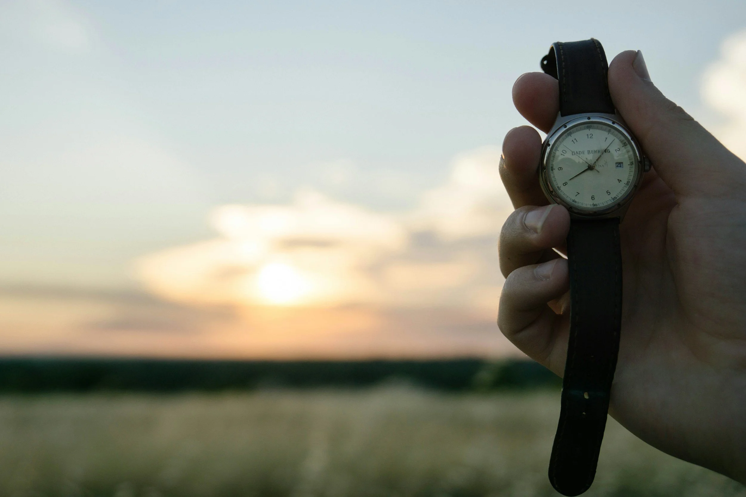 A hand holding a wristwatch with a sunset and open landscape in the background.
