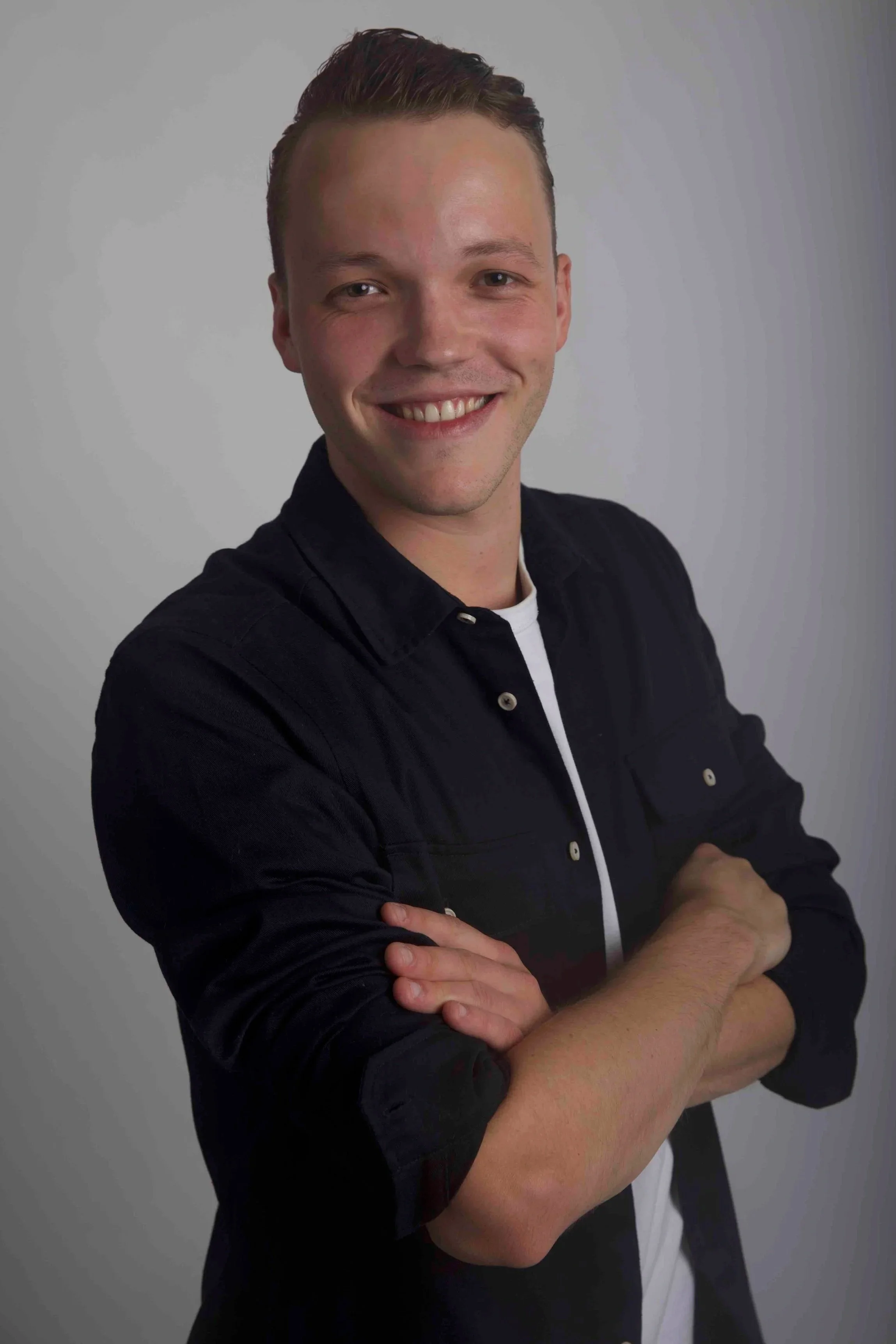 Portrait of a young man smiling with arms crossed, wearing a black shirt and a white t-shirt underneath, against a grey background.