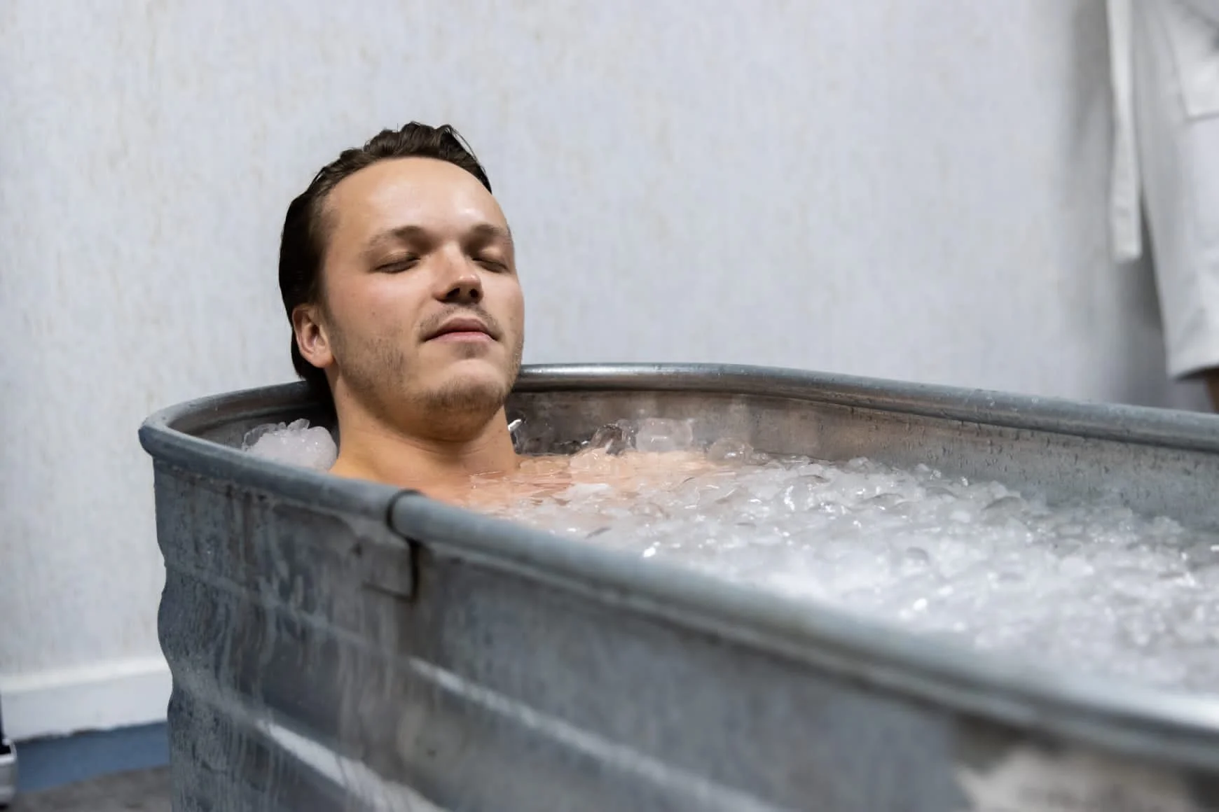 A man with closed eyes relaxing in a metal ice bath tub filled with ice water.