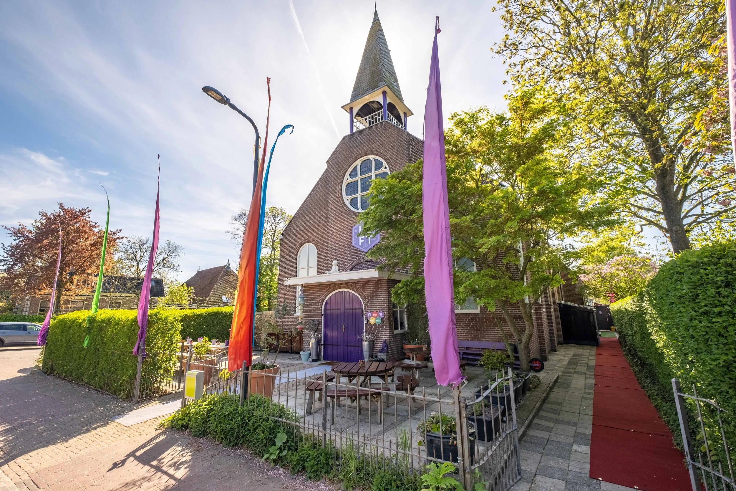 A brick church with a purple door and a tall bell tower, decorated with colorful flags and surrounded by green trees on a sunny day.