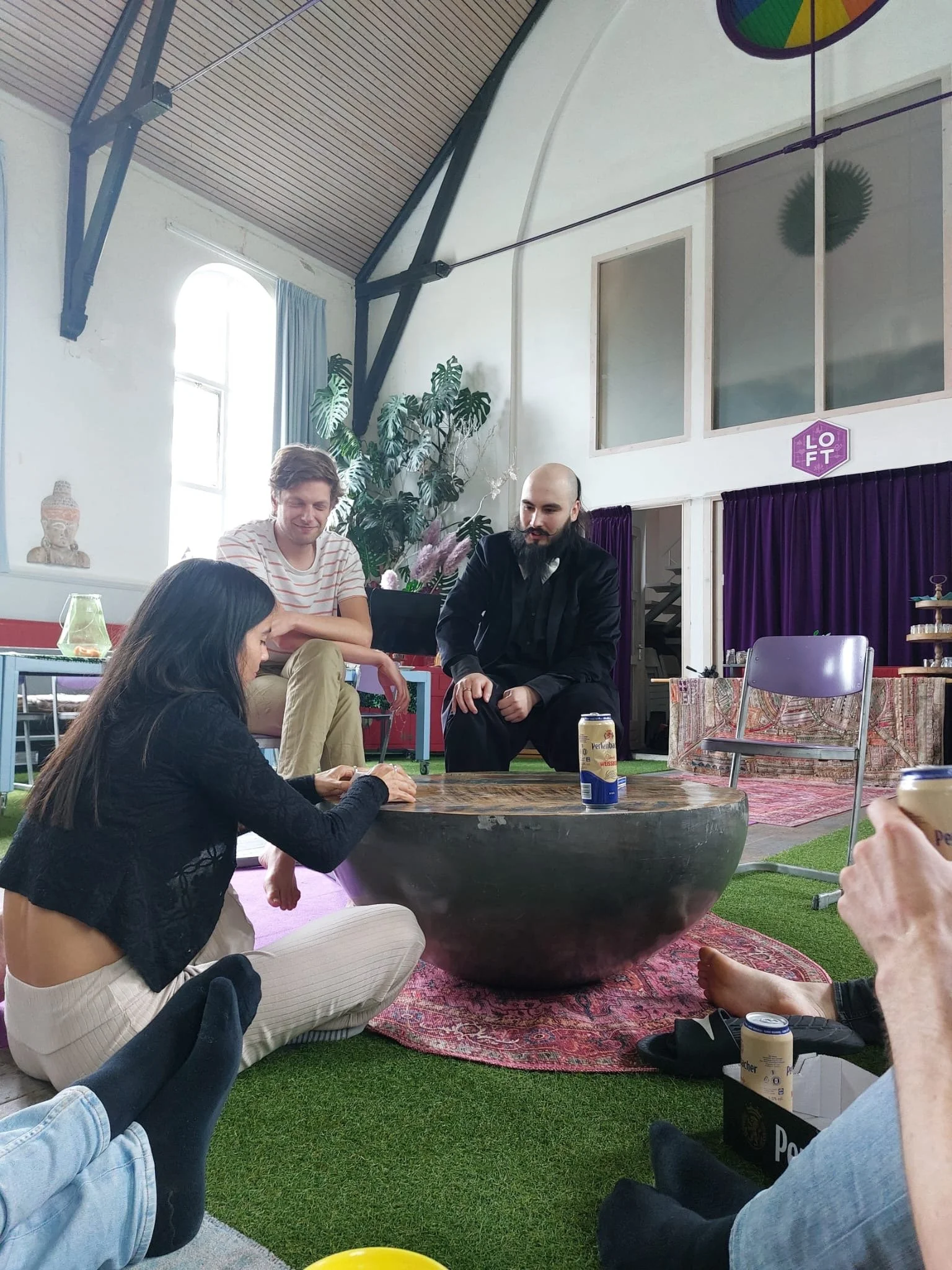 A group of friends sitting on the floor around a large, bowl-shaped table inside a bright, spacious room with high ceilings. They are enjoying drinks, with some holding cans. The room has large windows, plants, and colorful decor, creating a relaxed, social atmosphere.