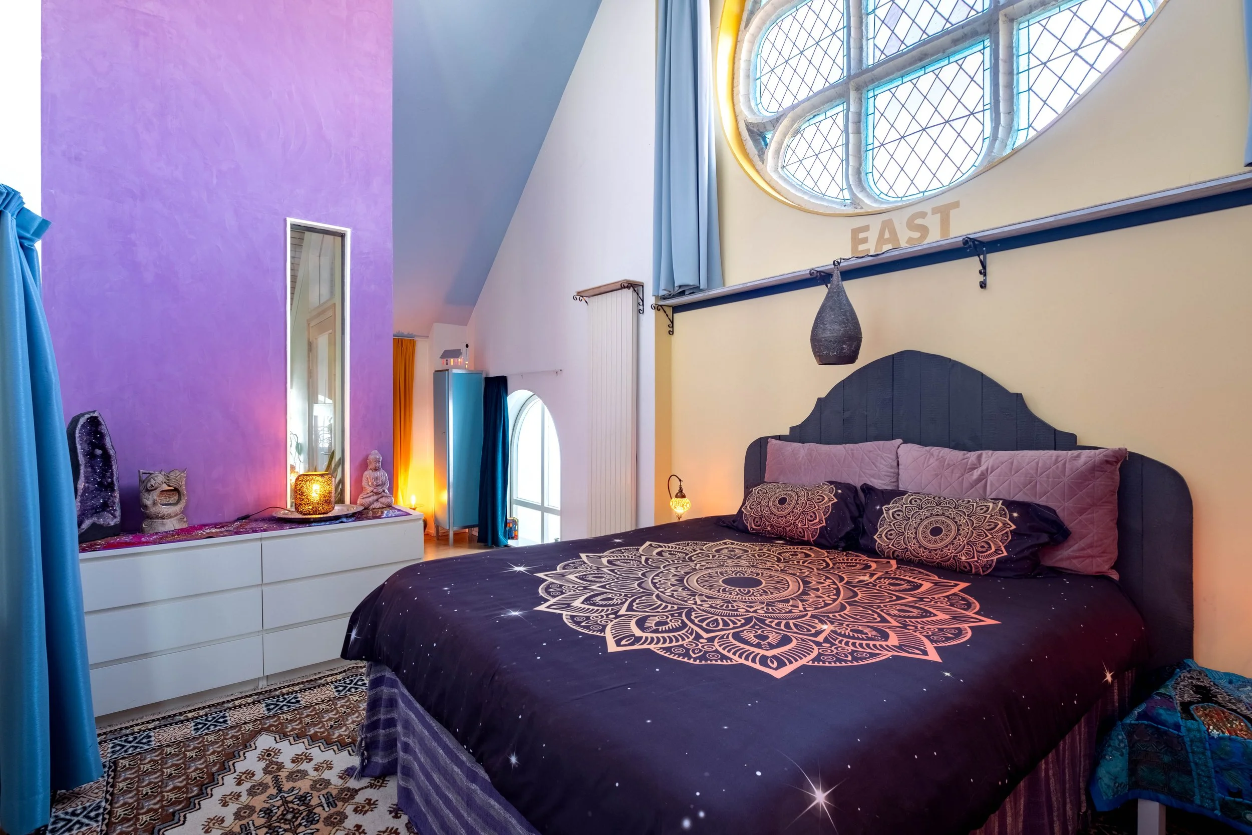 A colorful bedroom with a large bed that has a starry galaxy-themed bedspread and pillows. There are decorative objects on a white dresser, including a crystal and a Buddha statue. The room has a purple accent wall, a tall mirror, and a prominent stained glass window with the word 'EAST' beneath it. The space is decorated with warm lighting and various curtains.