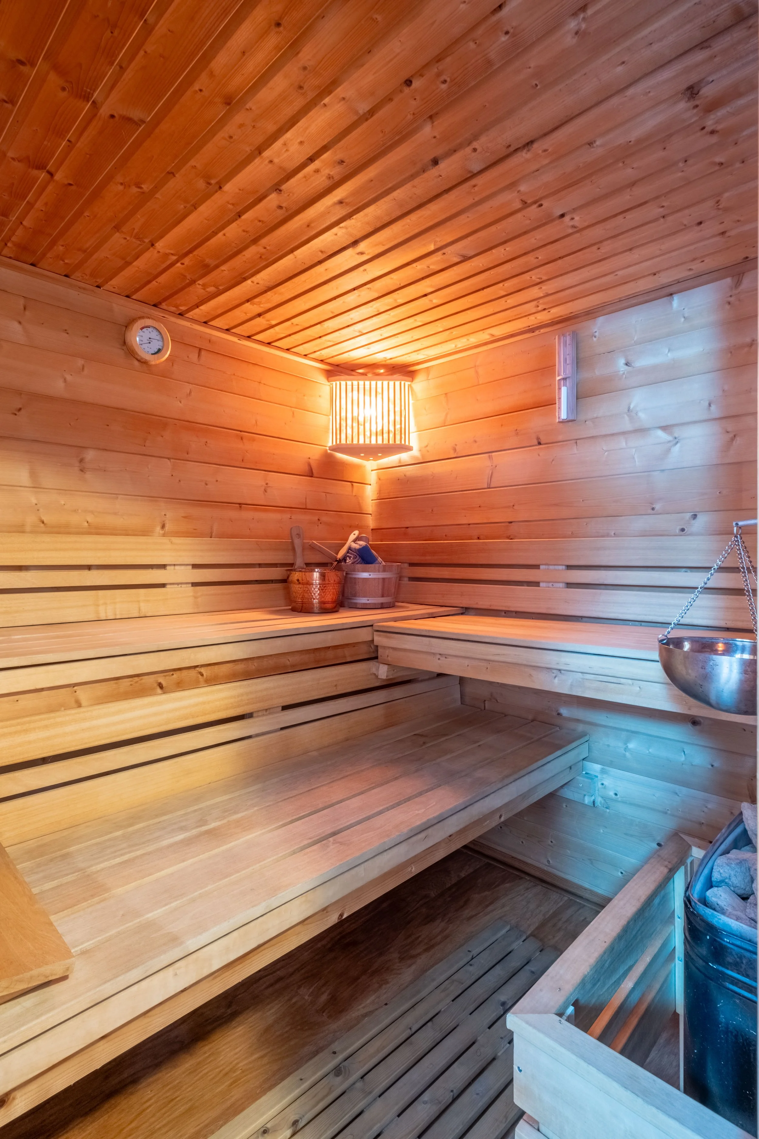 An interior view of a traditional wooden sauna with wooden benches, a bucket of water, a ladle, a thermometer, and a wall-mounted heater.