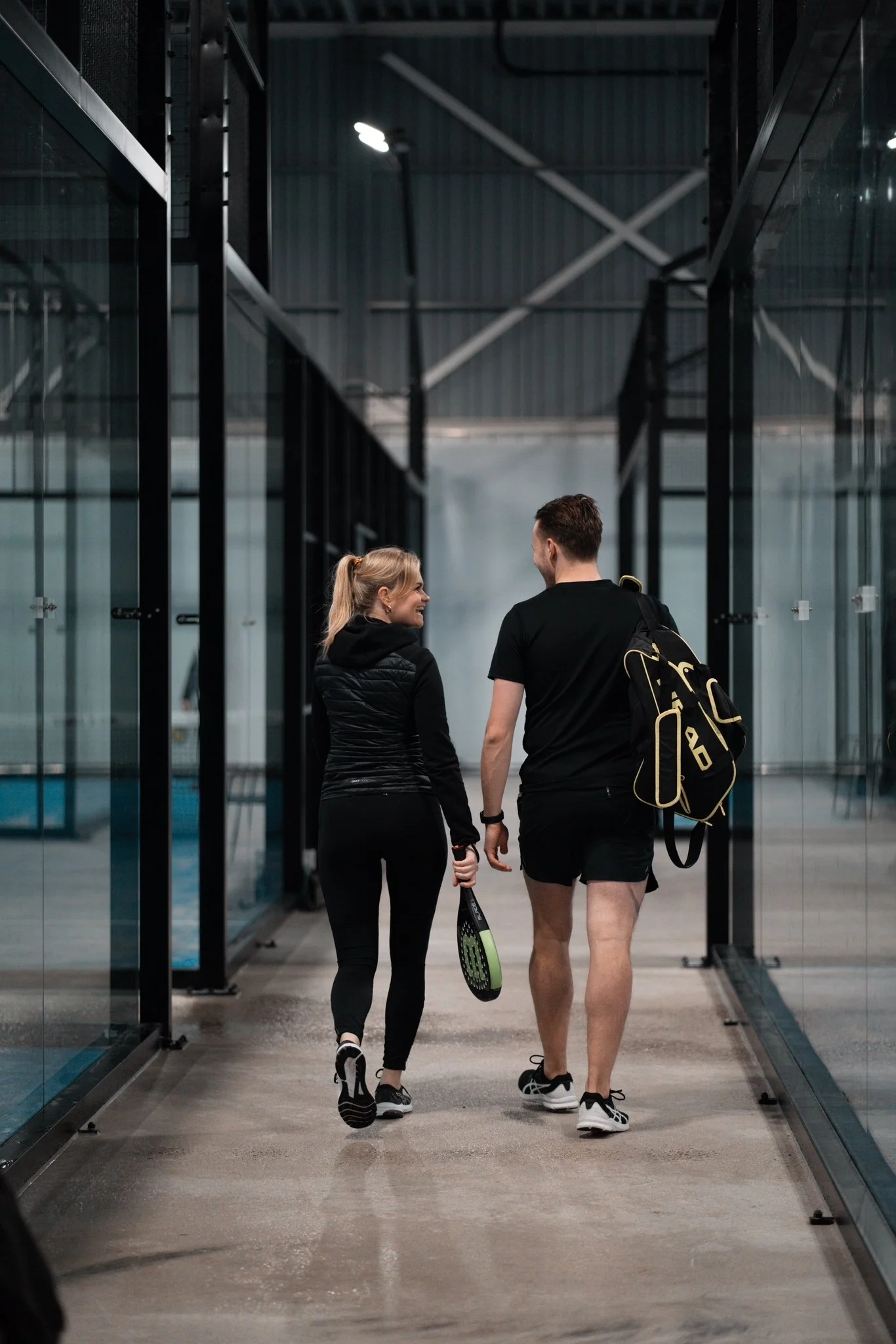 A man and woman walking together inside a modern indoor tennis facility, carrying tennis rackets and smiling.