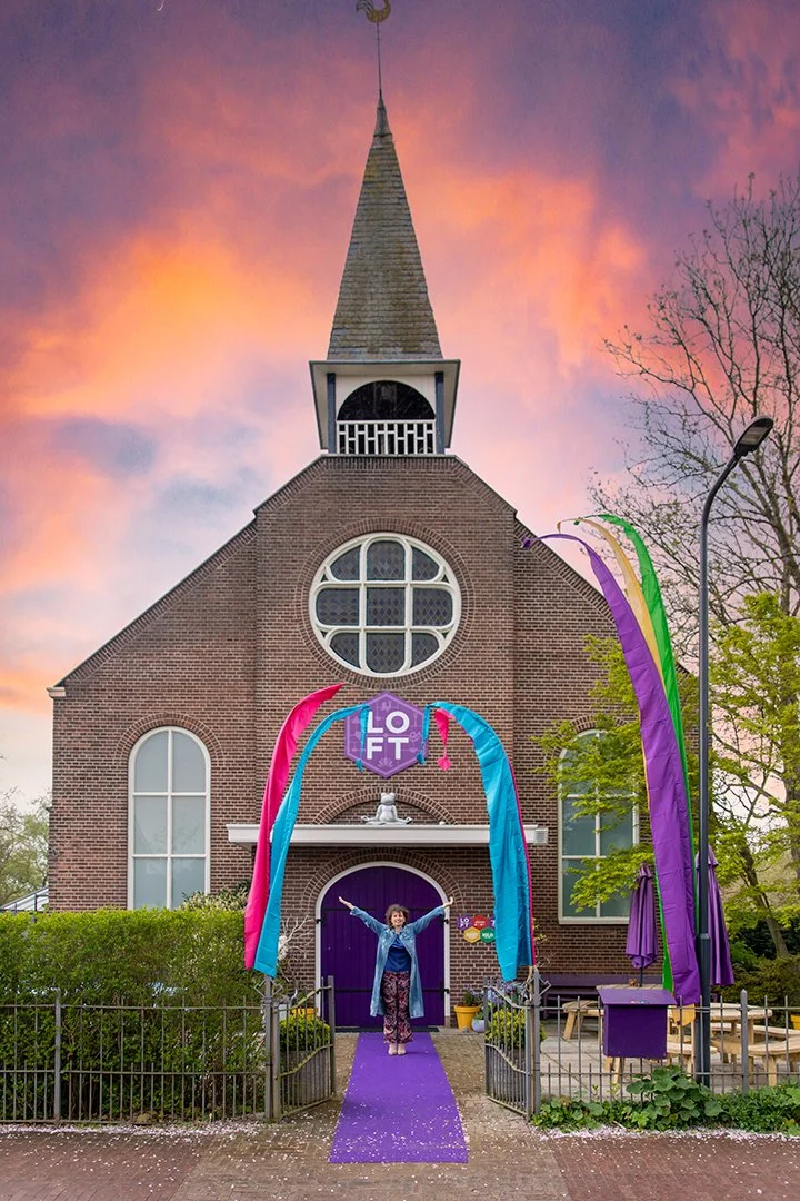 A person standing with arms raised in front of a small brick church decorated with colorful banners for a pride event, with a sunset sky in the background.