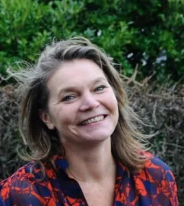 A woman with shoulder-length hair smiling outdoors with green foliage in the background, wearing a red and blue patterned top.