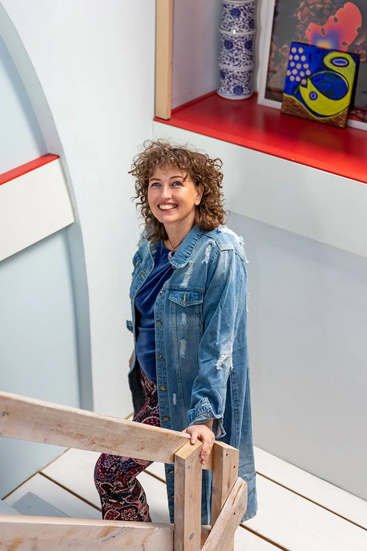 Woman with curly hair smiling and holding wooden railing in an art gallery or studio, with colorful artwork and blue patterned vase on a red shelf in the background.