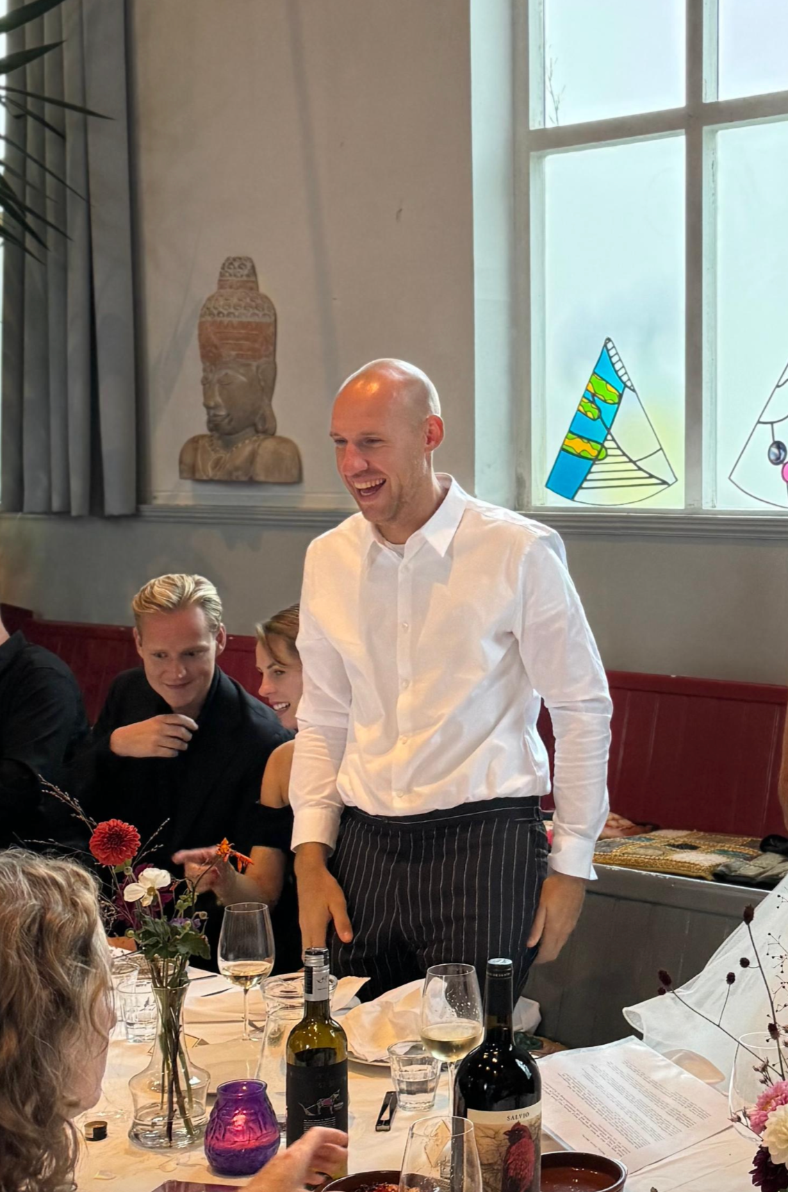 A group of people gathered around a dining table, with a man in a white shirt standing and smiling. The table has wine bottles, glasses, flowers, and plates. There are colorful drawings on the window and a sculpture on the wall.