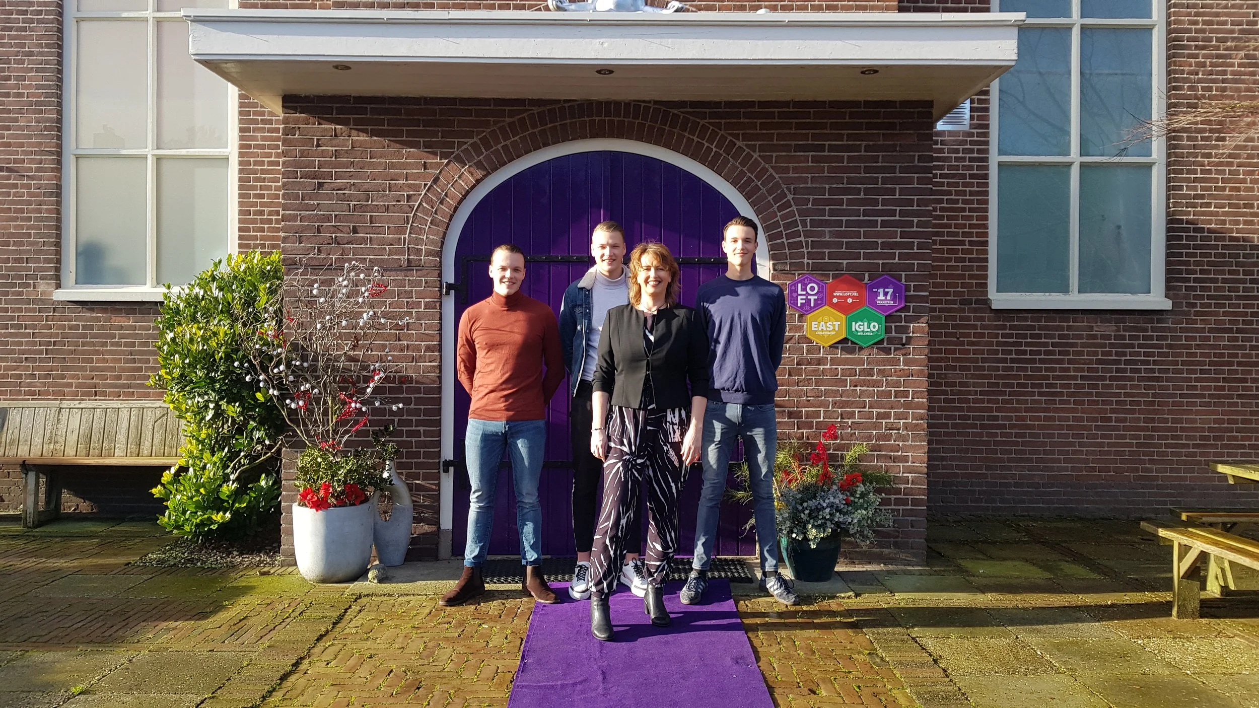 Group of four people standing in front of a brick building doorway with purple door and colorful signs, on a purple carpet with potted plants on either side.