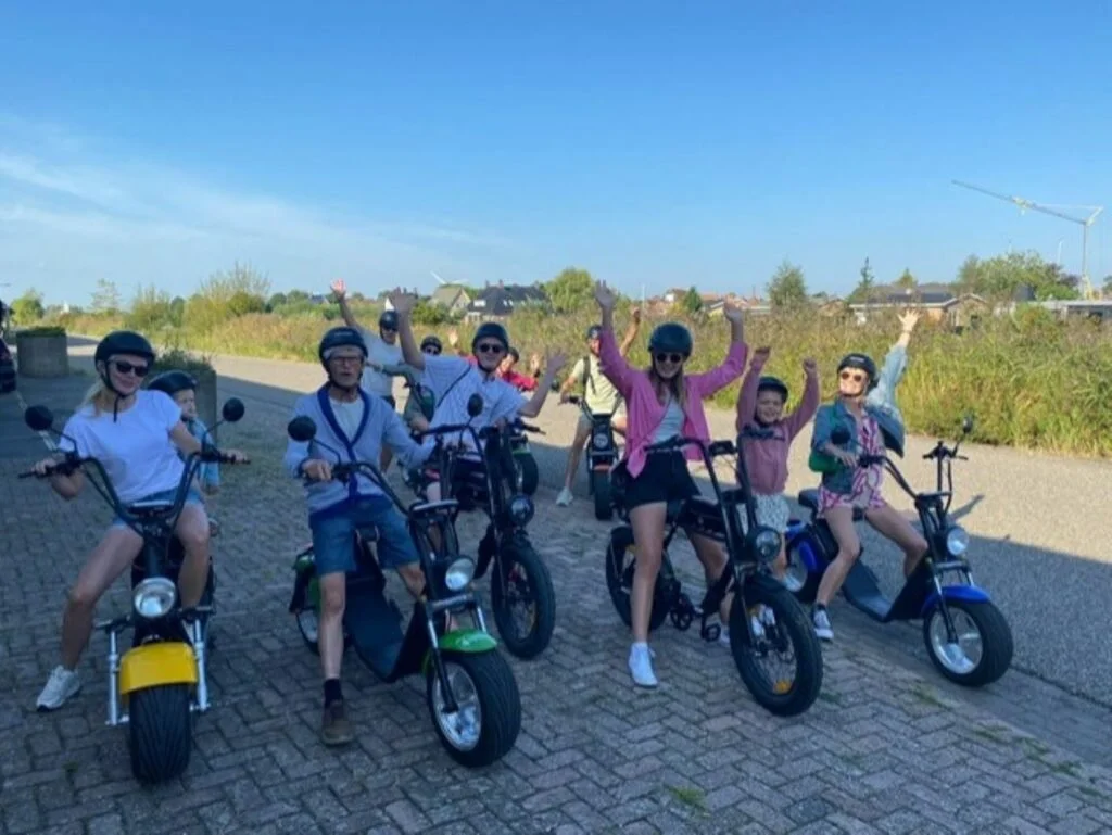 Group of people, including children, riding electric bikes outdoors on a clear, sunny day, smiling and raising their hands in the air.