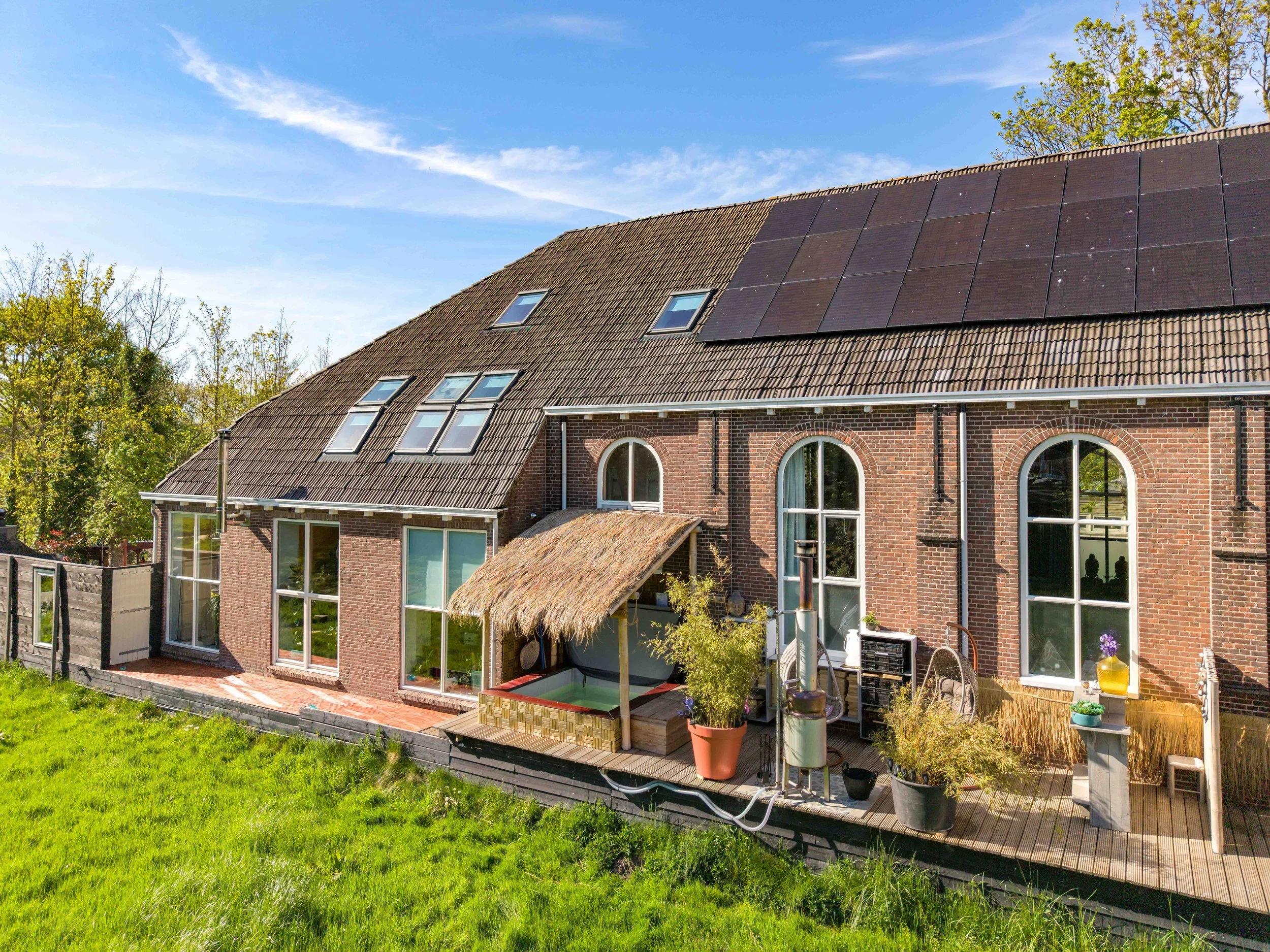 The back of a brick house with large arched windows and a sloped roof with solar panels and skylights. There is a wooden deck with potted plants, outdoor furniture, and a small thatched-roof structure.
