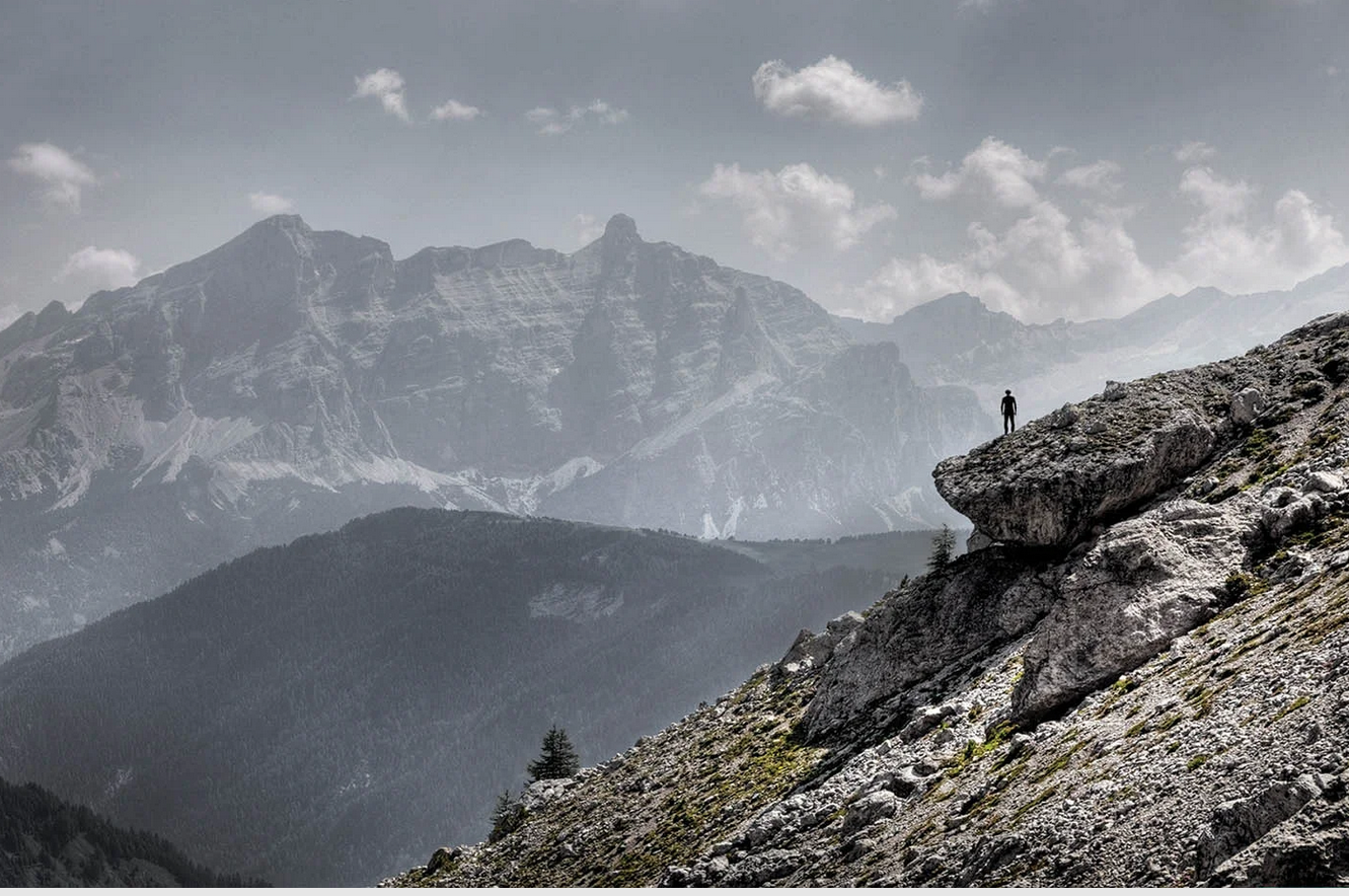 A person standing on a rocky mountain ledge overlooking a range of mountains under a partly cloudy sky.
