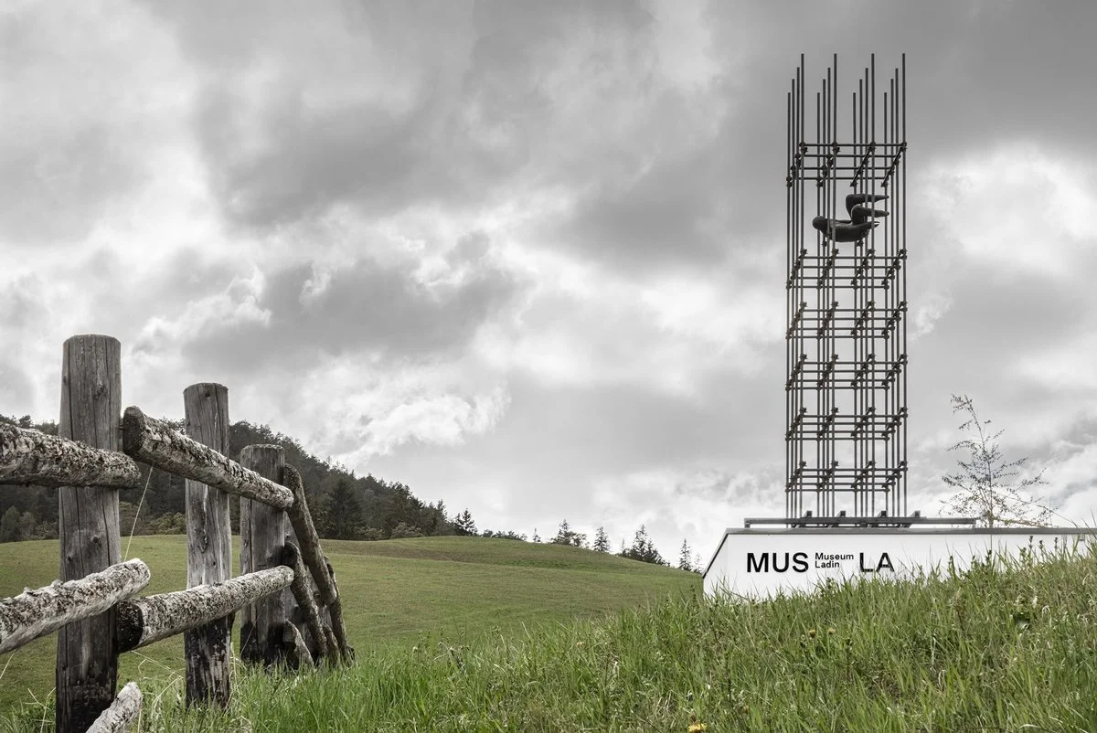 A metal scaffolding tower containing a dark, suspended figure stands on a grassy hill.