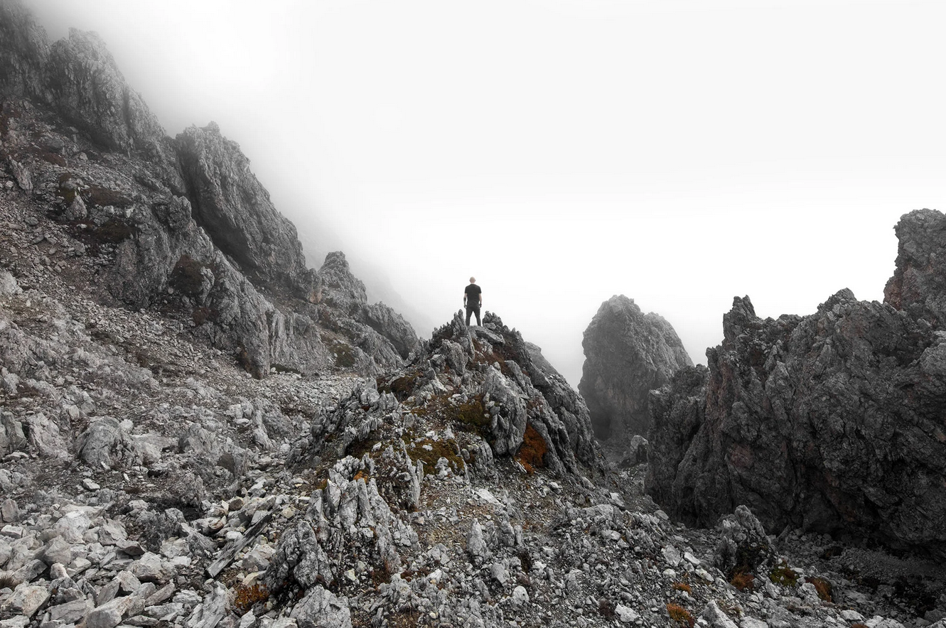 A person standing on a rocky mountain terrain with steep cliffs on either side, foggy sky overhead.
