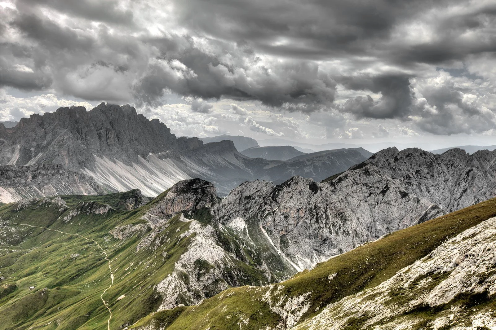 Mountain peaks and steep rocky slopes stretch across the horizon under a dramatic, heavy sky, with a narrow trail winding along the green ridges below.