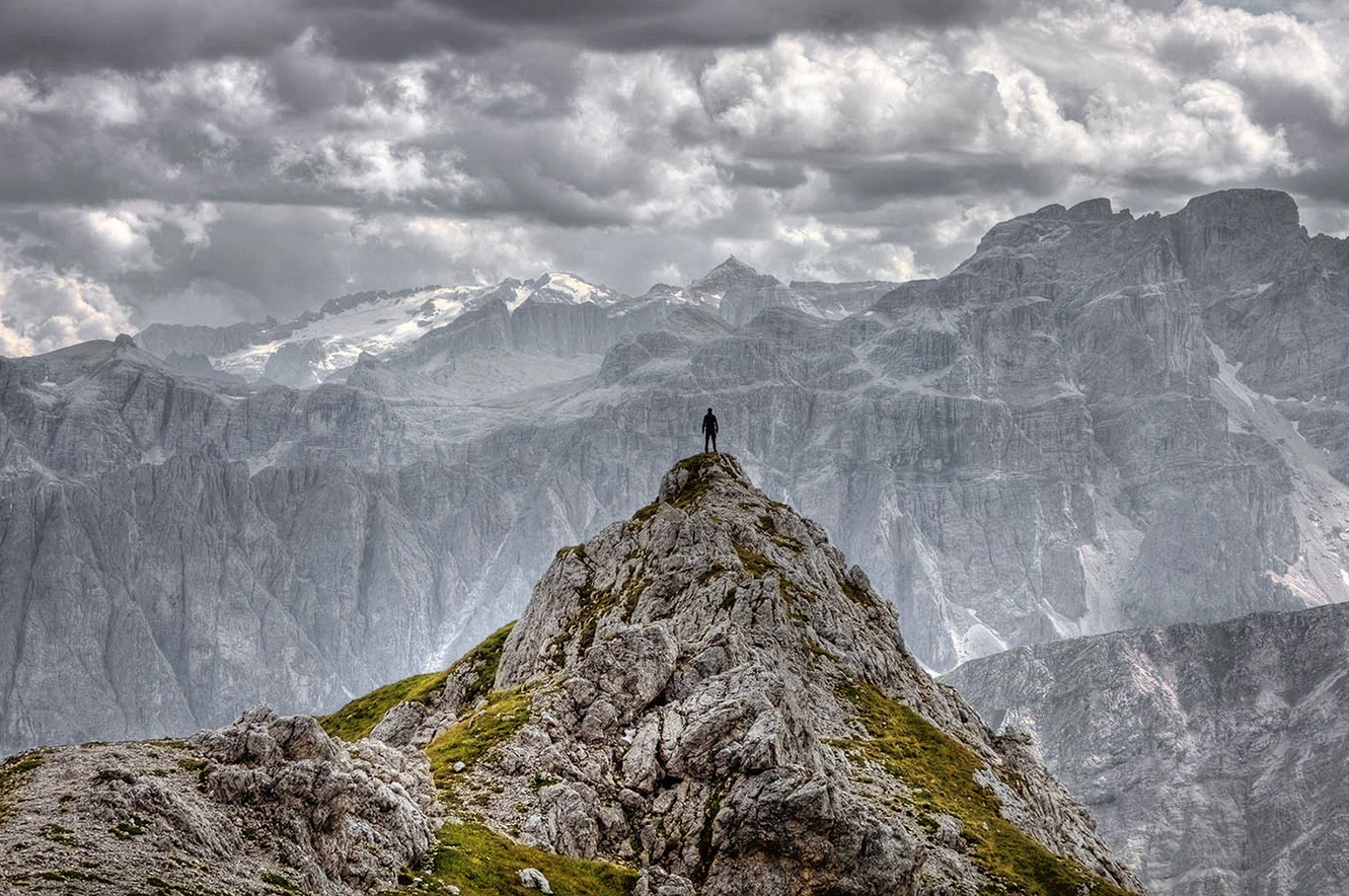 A person standing on a rocky mountain peak with a vast mountain range in the background and cloudy sky above.