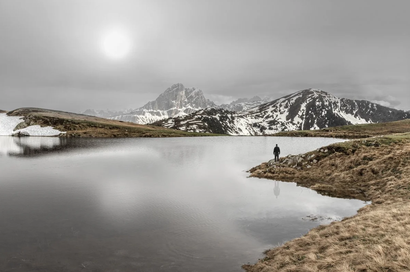 A person standing on the shore of a calm mountain lake, with snow-capped peaks and rugged hills in the background on a cloudy day.