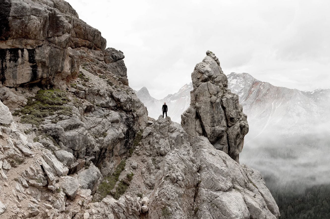 A human figure in black standing on a rugged rocky ridge, with towering mountain peaks and a misty valley in the background.