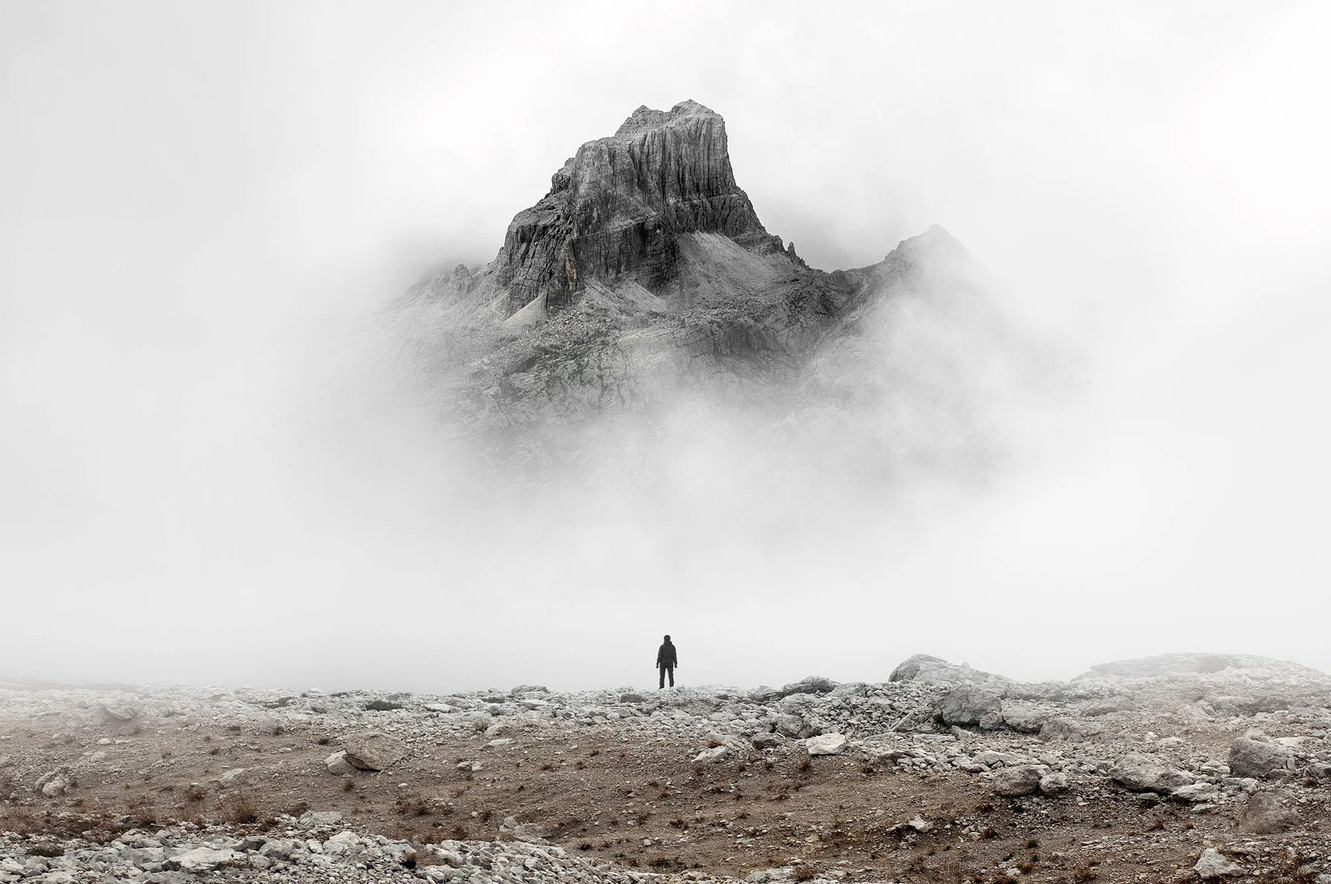 A person standing on rocky terrain in front of a mountain shrouded in fog.