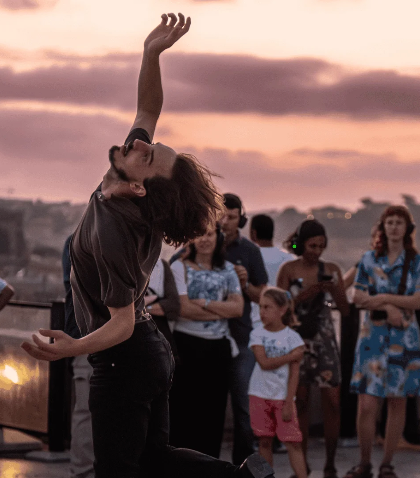 A male dancer performs an expressive, back-bending pose with one arm raised high on a rooftop at sunset, with an audience watching in the background.