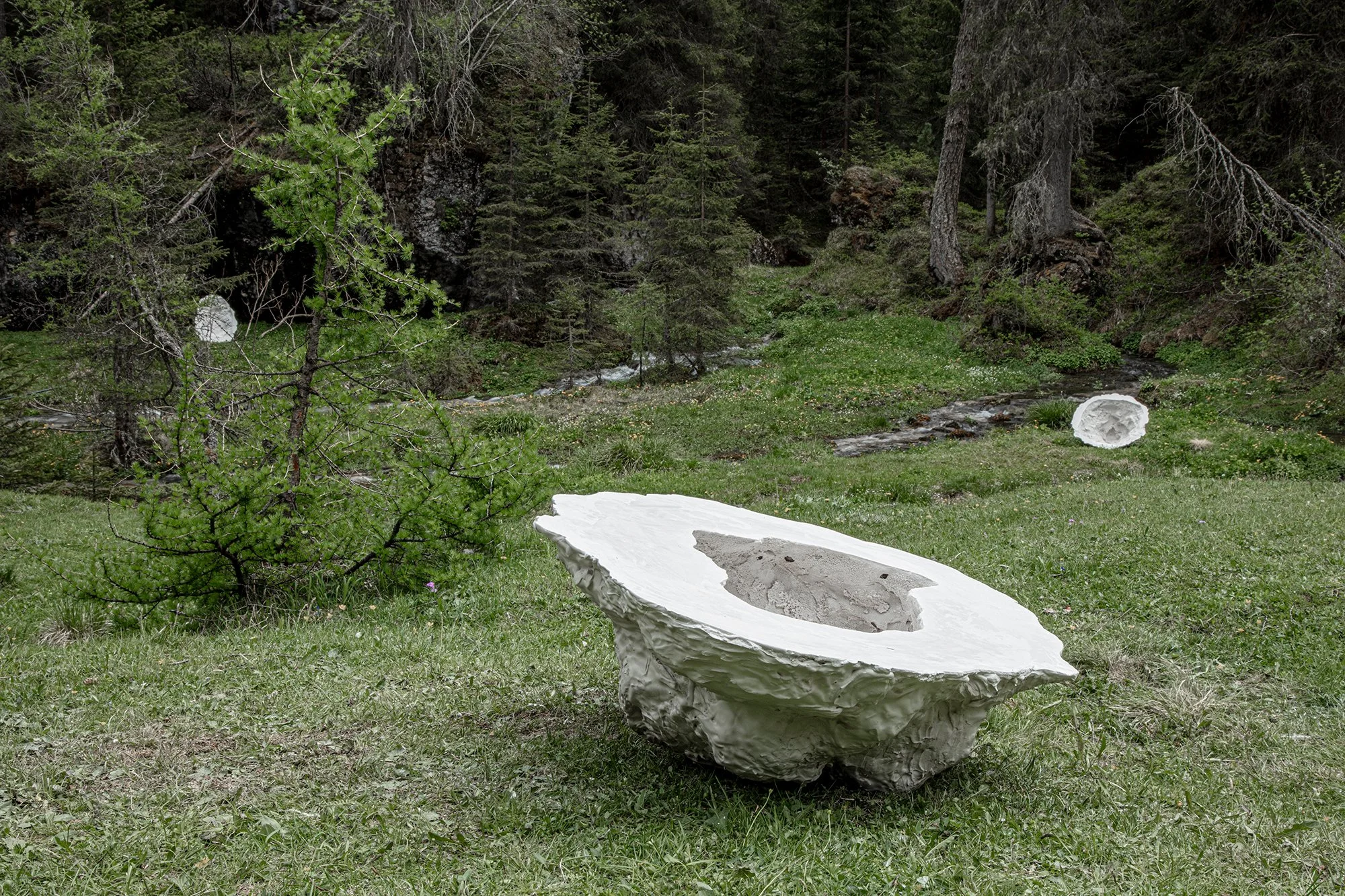 Three large, white, hollow organic-shaped sculptures scattered in a green meadow near a small stream and a forest.