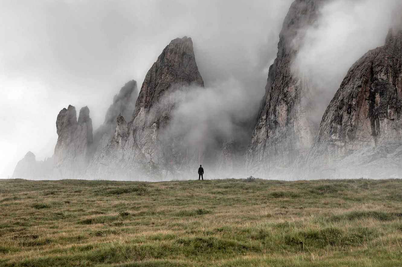 A person standing in a grassy field, with towering rocky mountains shrouded in fog and mist in the background.