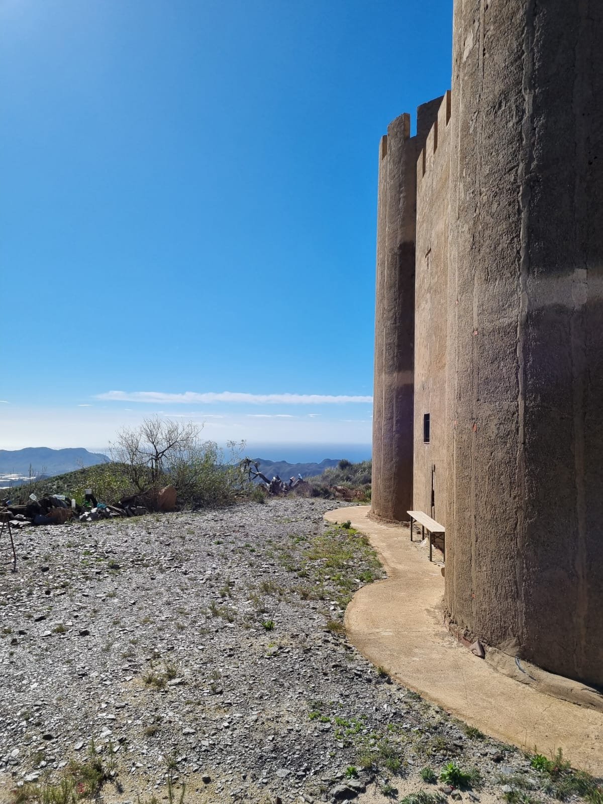 Ein historisches, zylindrisches Gebäude mit Mauern aus Lehm oder Ziegeln steht an einem würzigen, trockenen Gelände. Es gibt eine Bank vor dem Gebäude, wo man die Aussicht auf die Berge und das Meer im Hintergrund genießen kann.