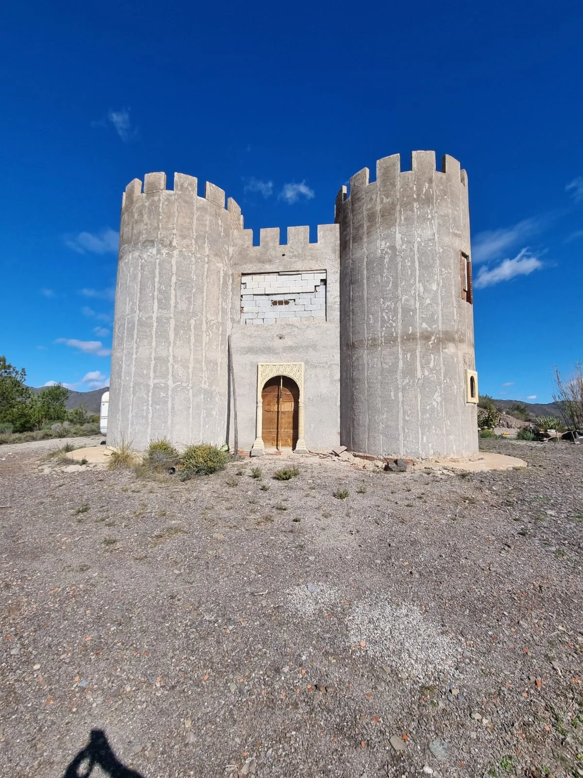 Ein fertiges, graues Schloss mit Tür und kleinen Fenstern, auf einem Kiesplatz mit wenigen Pflanzen, unter blauem Himmel mit einigen Wolken.