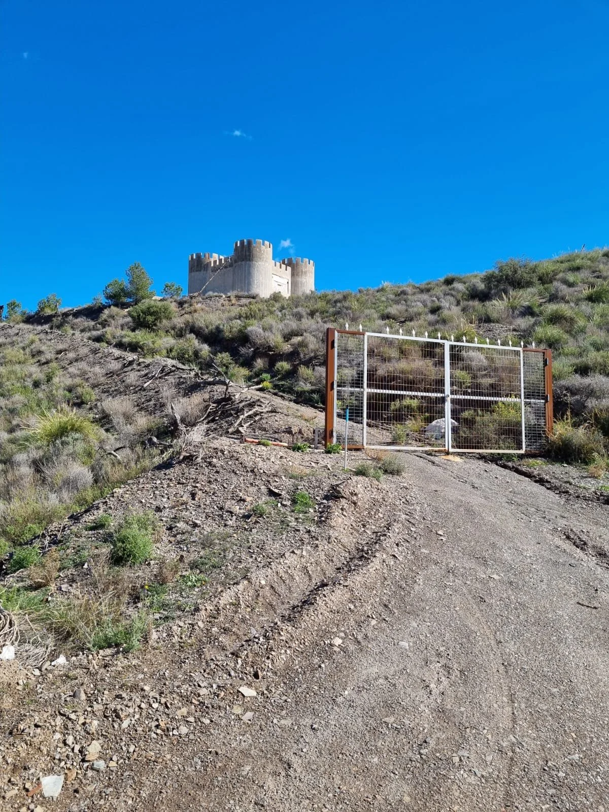 Ein steiniger Weg führt zu einem Zaun und einem Schloss auf einem Hügel unter einem klaren blauen Himmel.