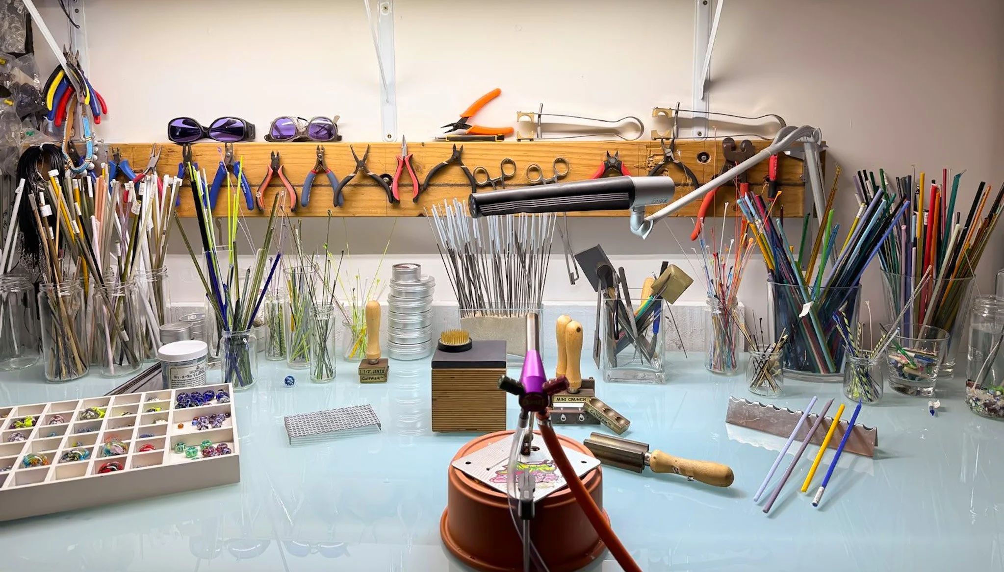 Jewelry making workspace with tools, beads, and colorful wire organized in jars and containers, with a workbench and a wooden pegboard holding pliers and sunglasses.