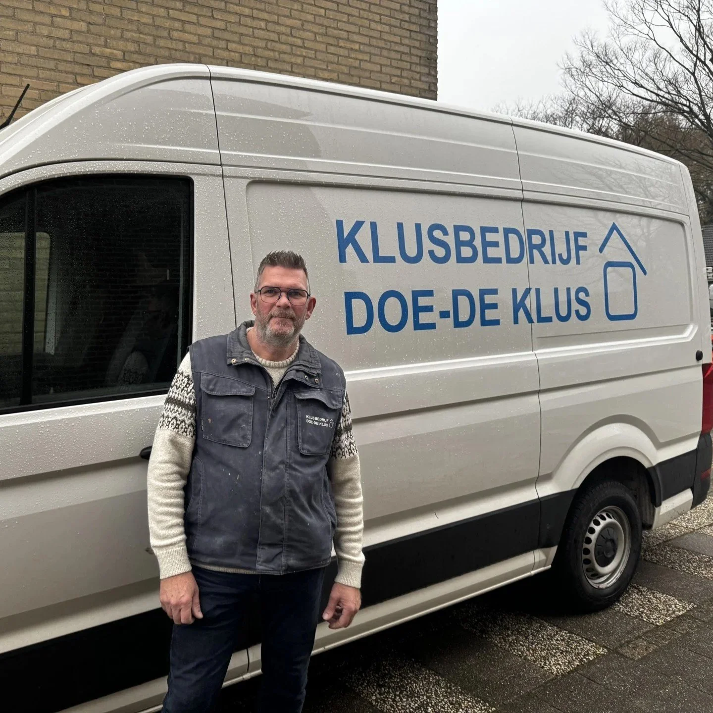 A man standing next to a white van with blue text that reads 'Klusbedrijf Doe-de Klus' on the side, outside in a cloudy setting with trees in the background.