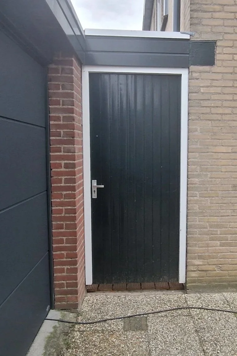 A black door with a silver handle set between a brick wall and a gray panel wall, with a concrete and brick step at the bottom and a black wire on the ground.
