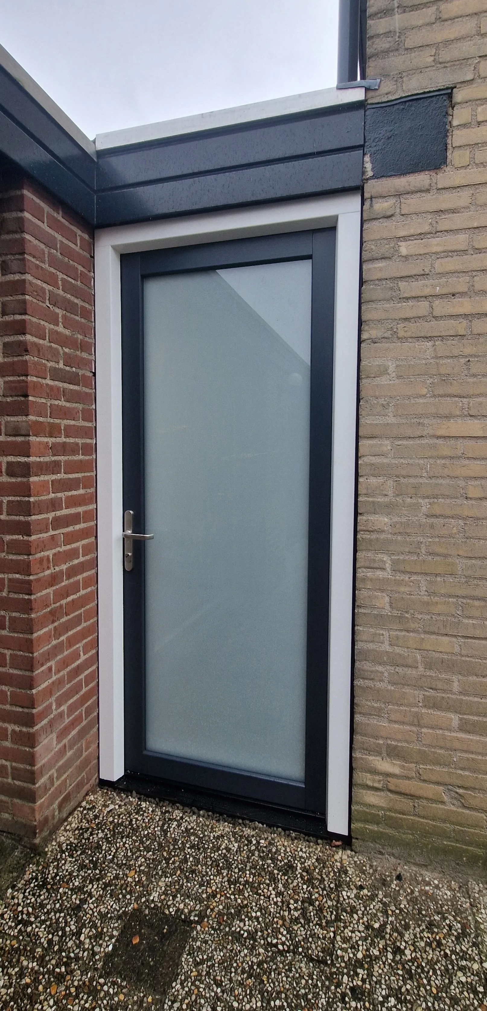 Exterior door with frosted glass pane, black frame, and silver handle, set between brick walls with gravel ground.